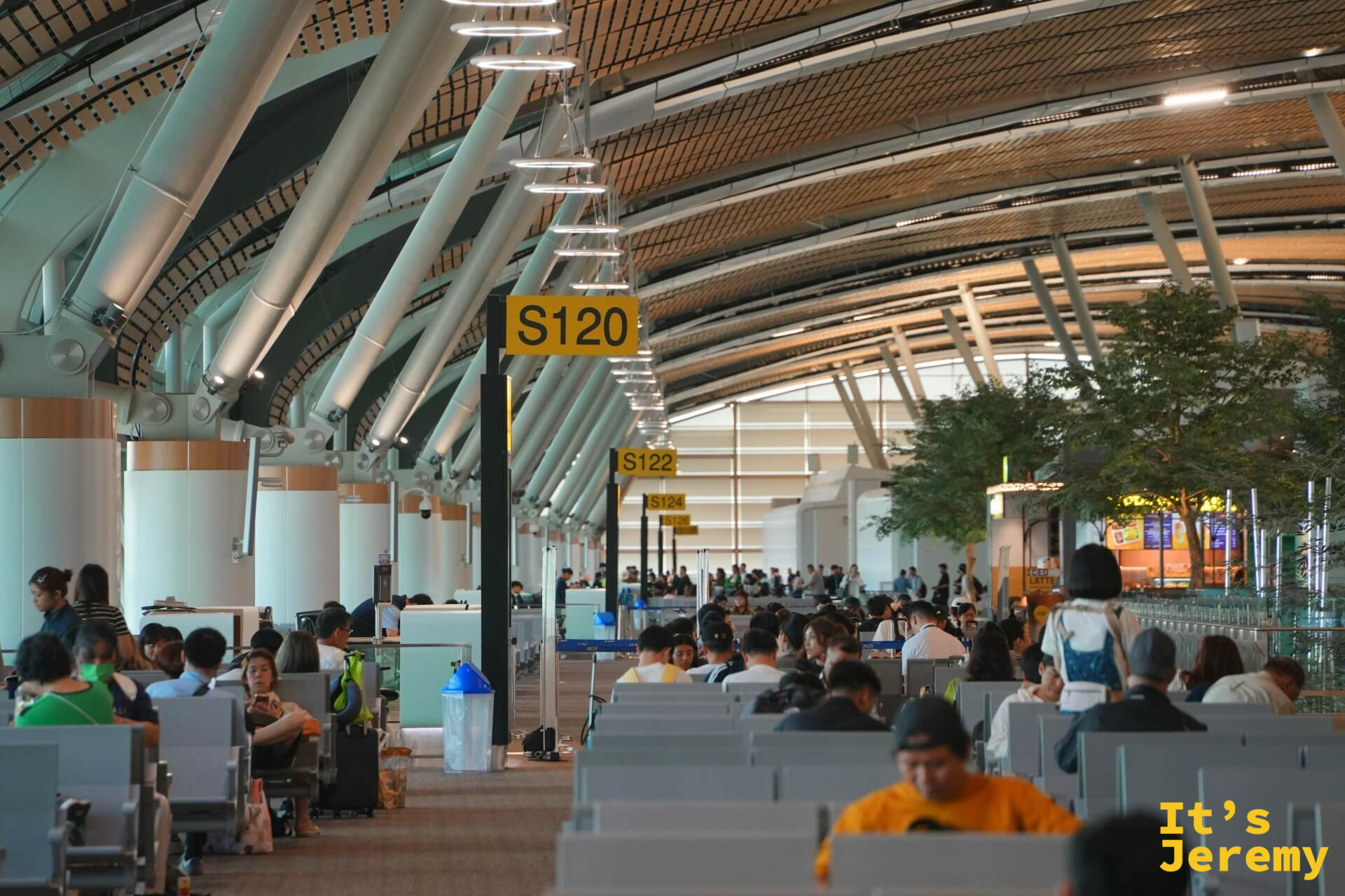 素萬那普機場 S120 候機室,木色弧形天花板,排滿候機旅客,S122 標示牌,旁邊有植栽
