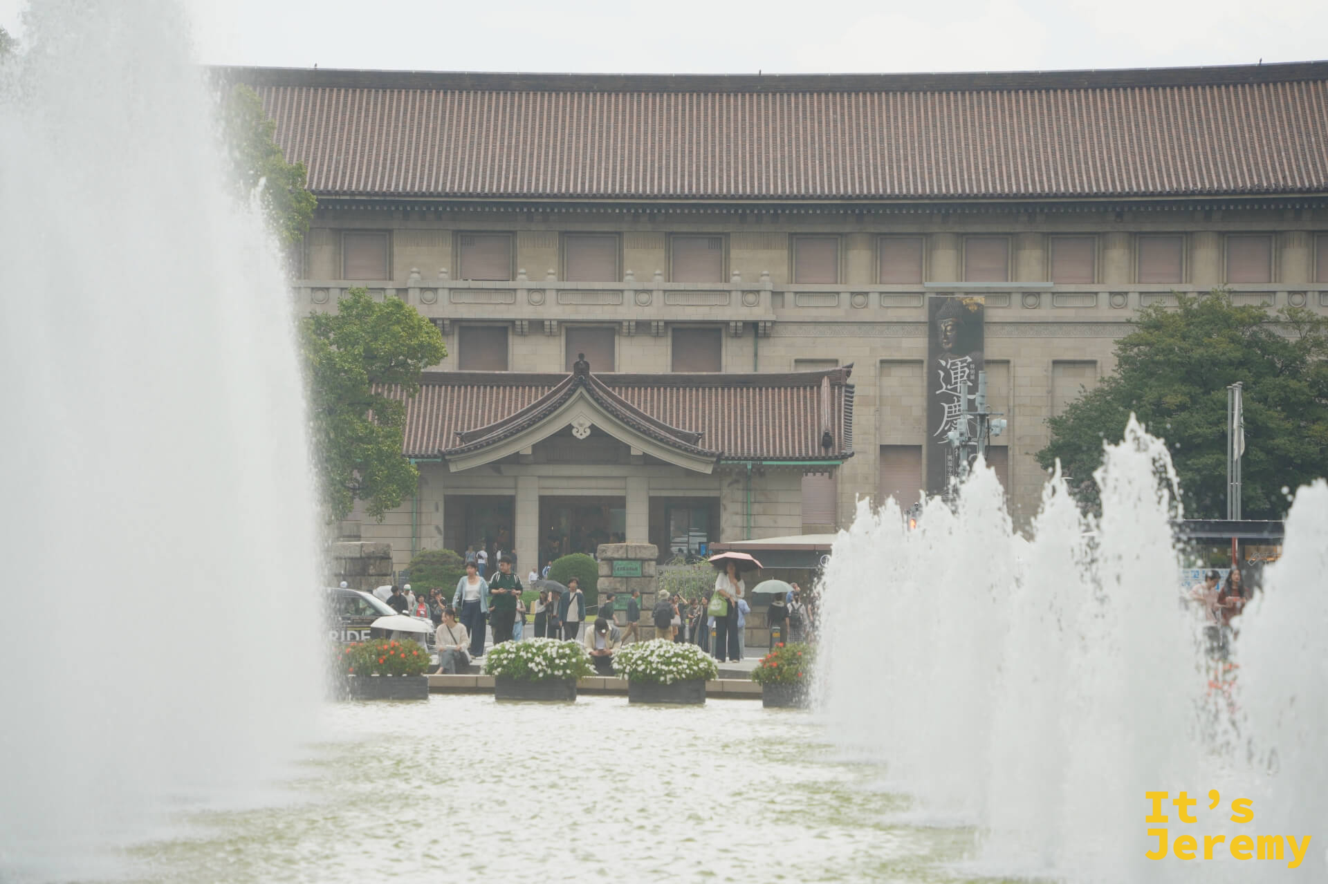 透過噴水池的水霧，東京國立博物館正面建築的日式屋頂清晰可見，廣場前有撐傘的遊客散步其中