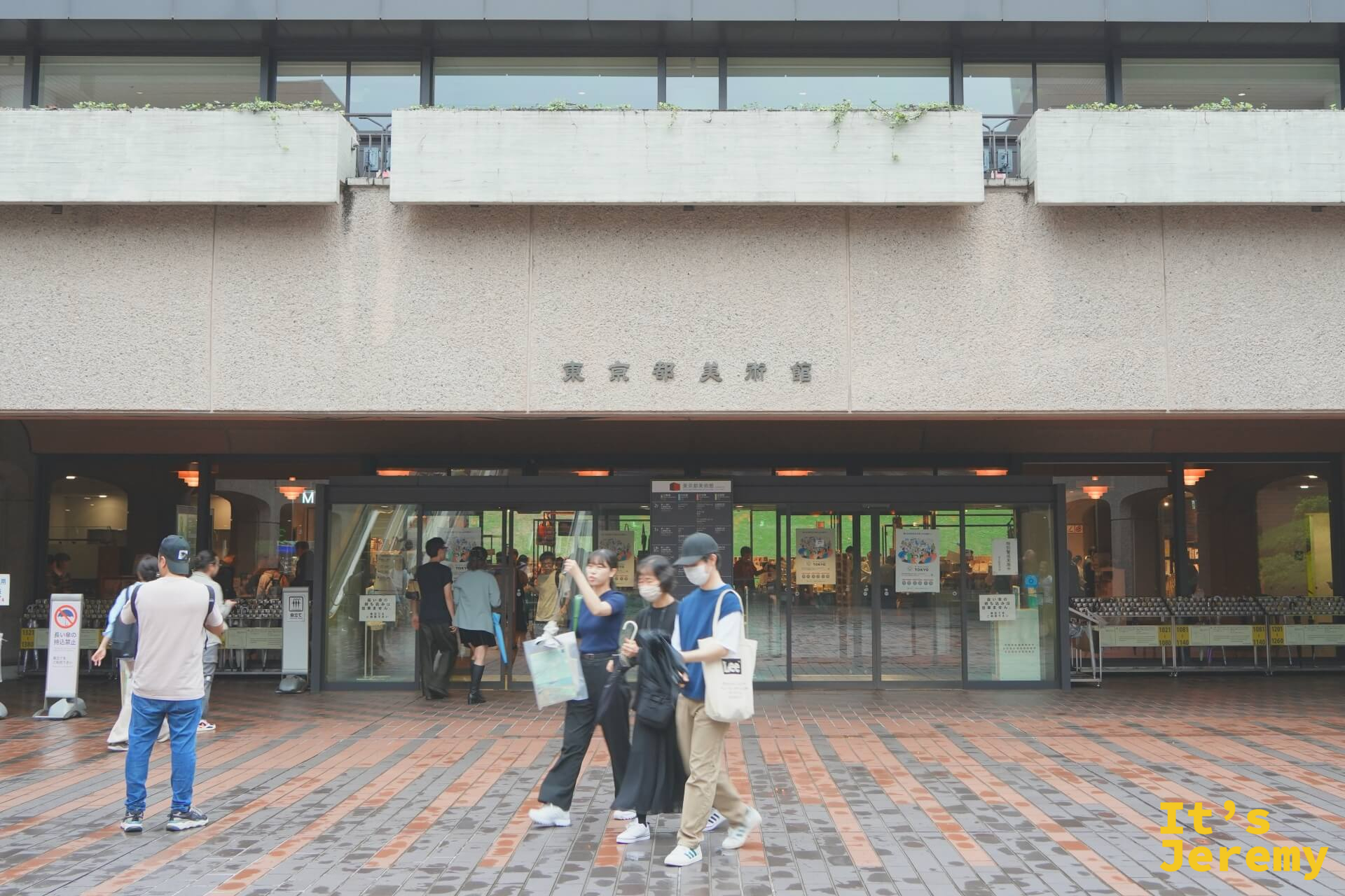 東京都美術館正面入口廣場，磚石地面，「東京都美術館」字樣清晰可見，幾位年輕人從玻璃門魚貫走出