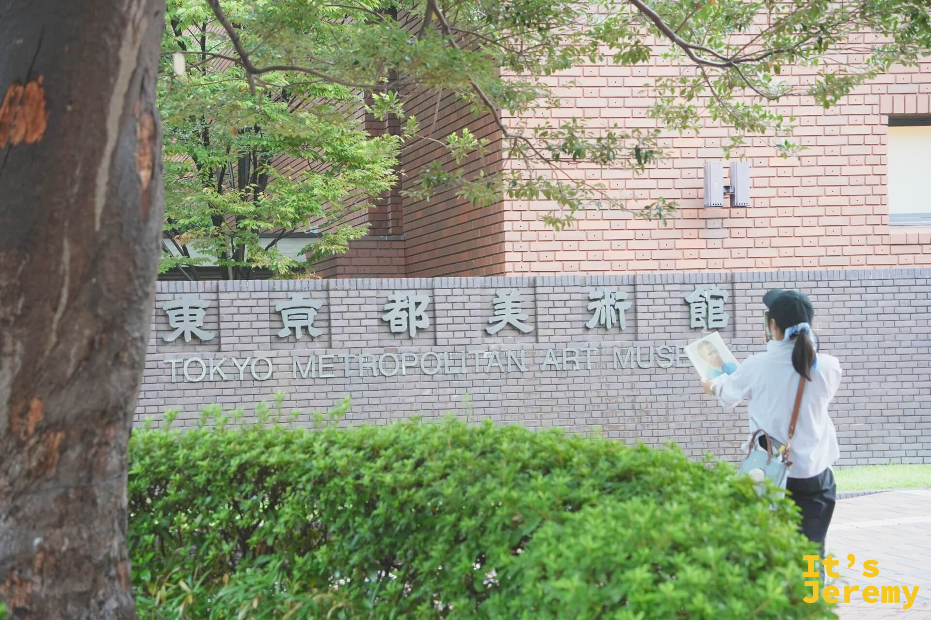 一位女士手持手冊站在東京都美術館外的標示牌旁，標示牌上有「東京都美術館 TOKYO METROPOLITAN ART MUSEUM」的中英文字樣，周圍是茂密的綠樹