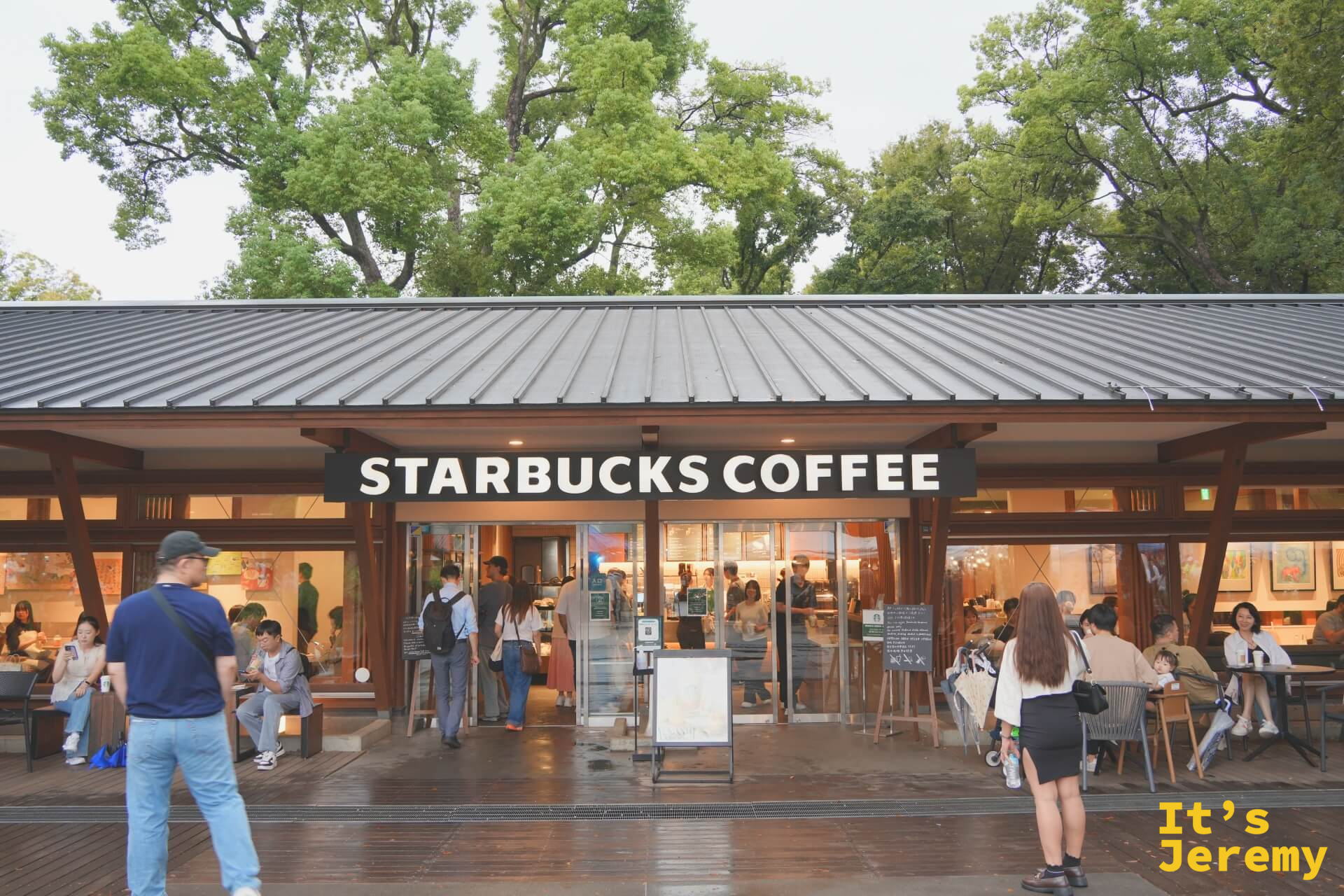 上野公園的星巴克全景，低矮木屋頂建築，「STARBUCKS COFFEE」招牌清晰，戶外座位坐著客人，背後是公園高大的老樹