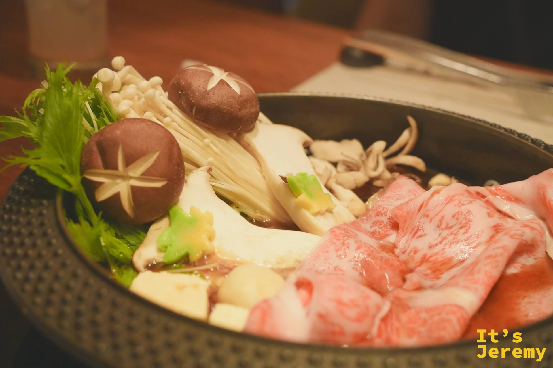 飛騨牛壽喜燒鍋端上桌，霜降肉捲與菇類蔬菜在黑色鑄鐵鍋中，食材尚未下鍋，霜降紋路在暖燈光下清晰可見