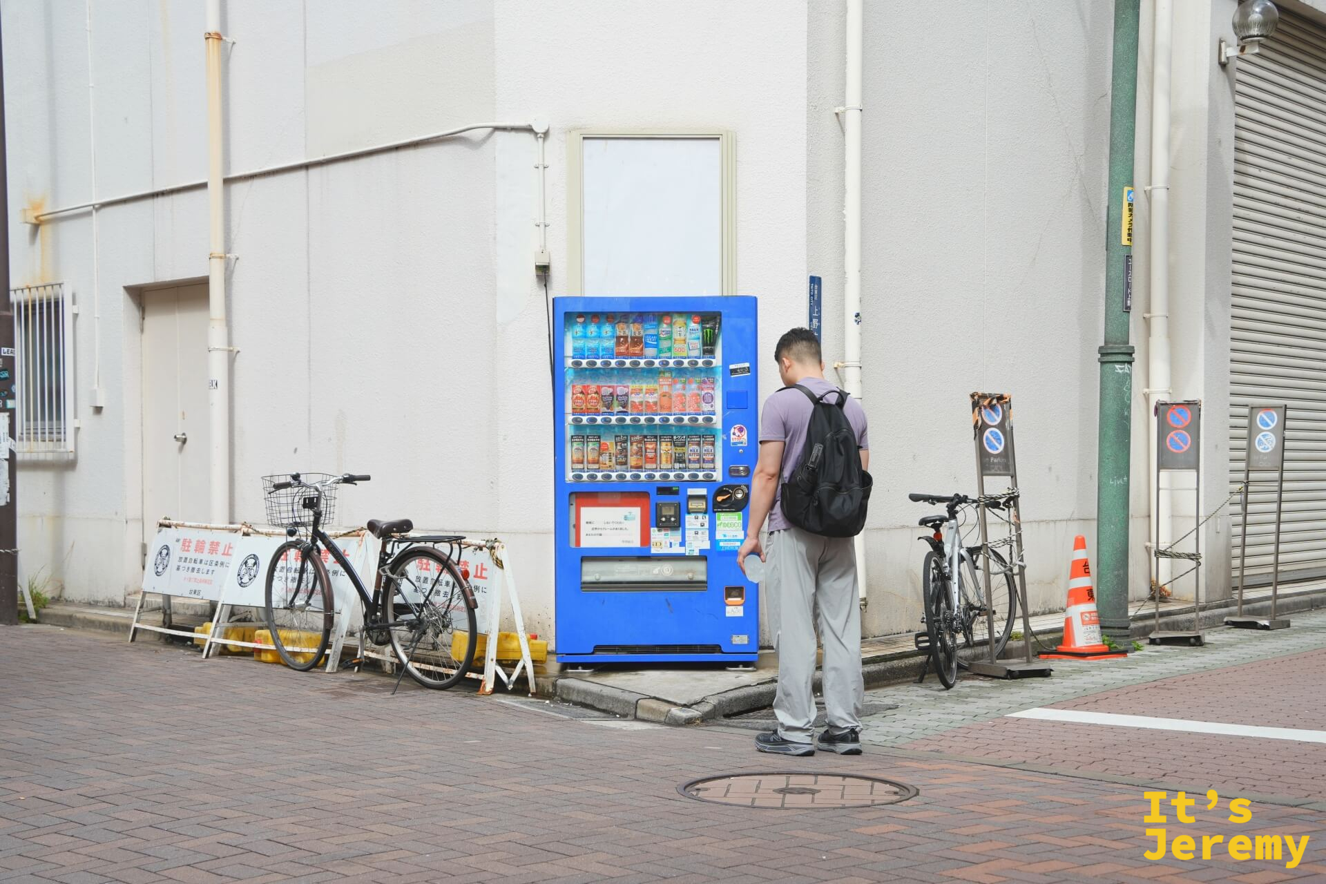 一個路人背對鏡頭站在藍色自動販賣機前選飲料,旁邊停著幾台腳踏車,是一個普通的東京街角