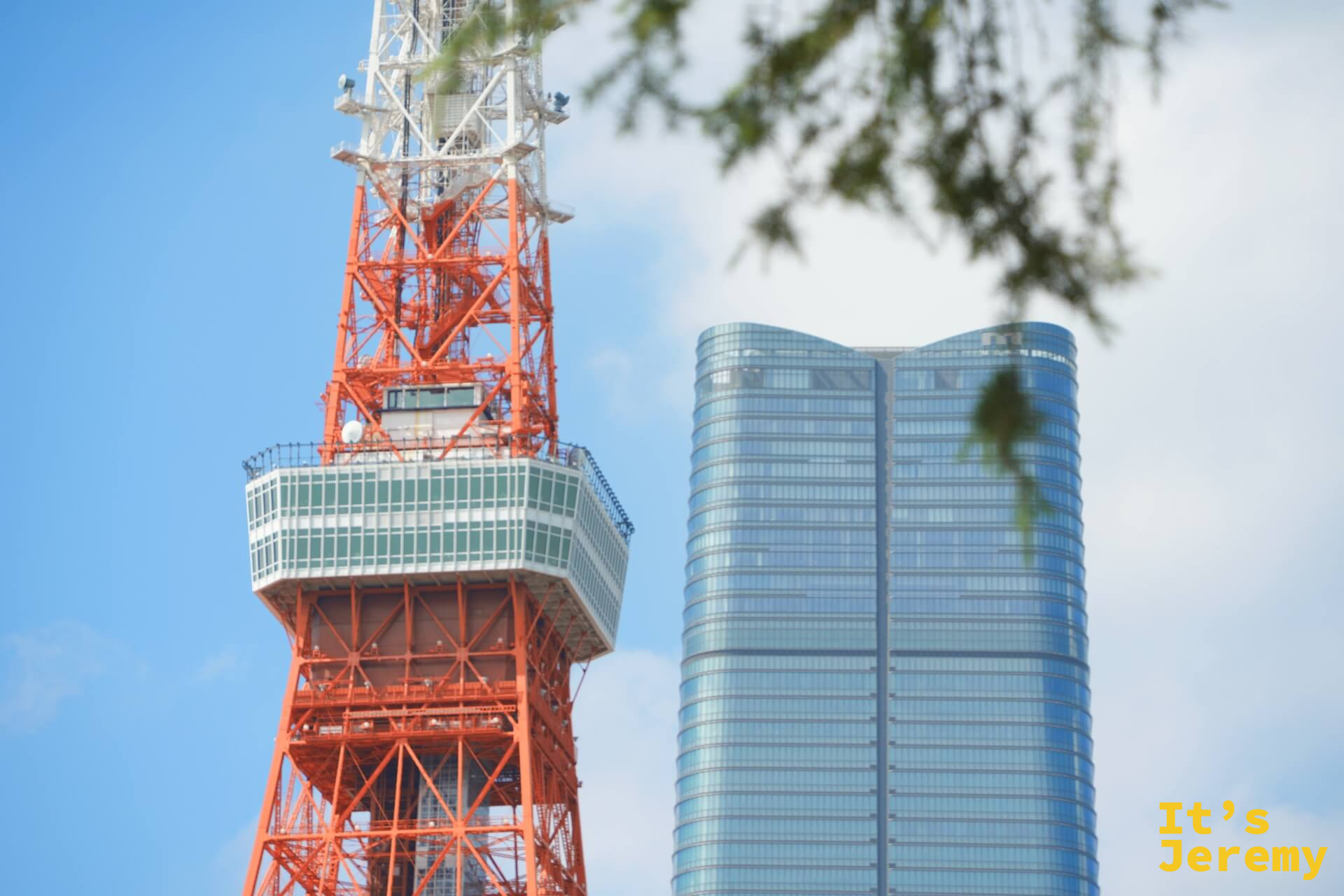 東京鐵塔的大展望台與旁邊一幢弧形玻璃帷幕高樓並排入鏡，鐵塔的橘紅格架與現代建築的冷灰玻璃形成強烈對比，前景有樹枝入鏡
