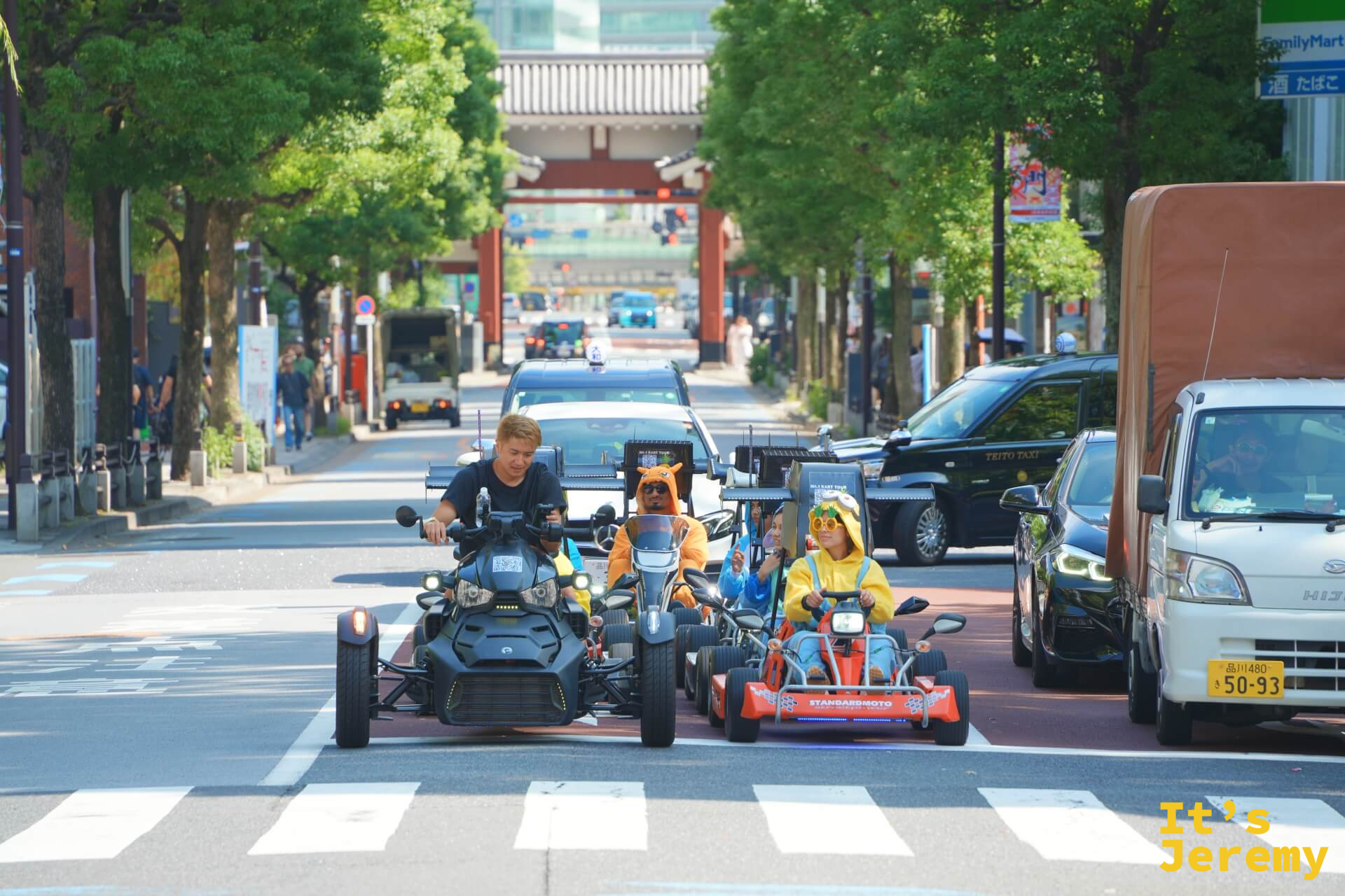 一列卡丁車正駛過芝大門路口,駕駛們穿著動漫角色服裝,後方可見一座朱紅色的鳥居式建築,普通車輛在兩側夾道