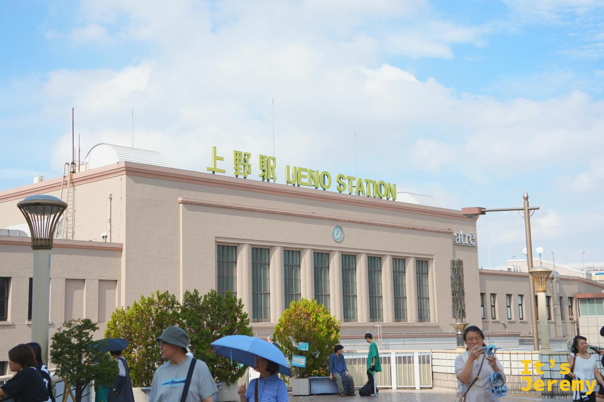 JR 上野駅外觀,「上野駅 UENO STATION」綠色大字頂在米色建築上,藍天白雲,前景是來往的旅客