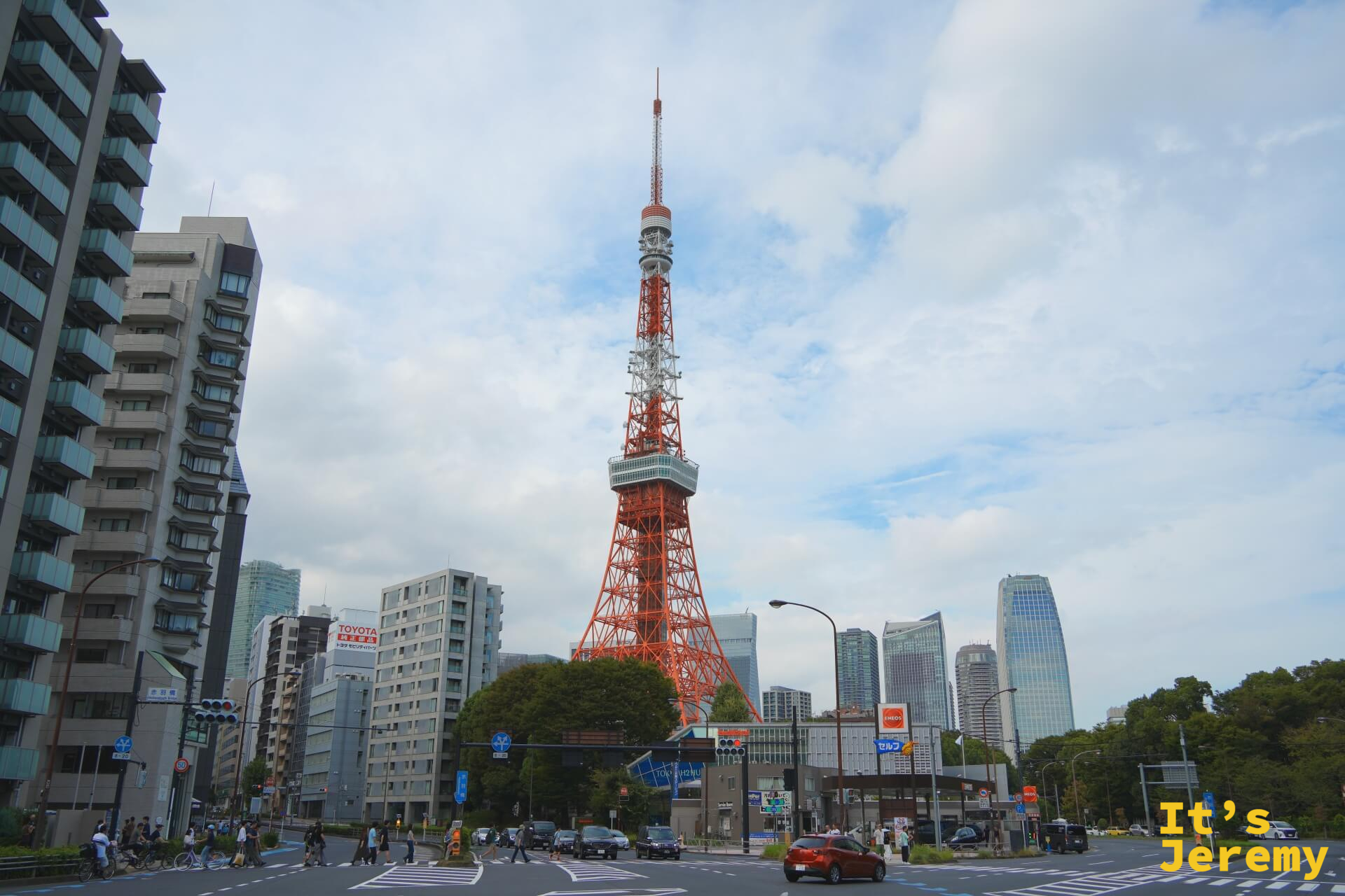 從赤羽橋交叉路口望向東京鐵塔，鐵塔矗立在城市建築群中央，橘紅色的鐵架在白色天空下格外搶眼，前景是普通的都市路口與行駛中的車輛