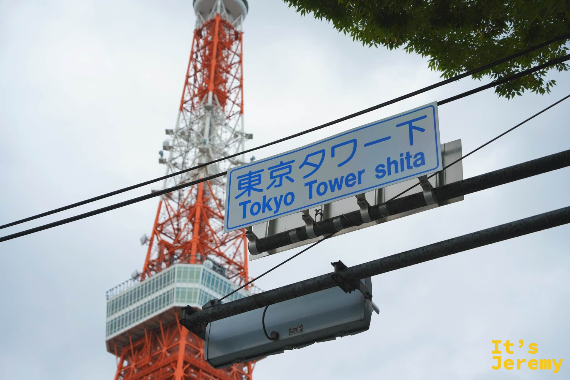路口電線桿上的藍白路牌寫著「東京タワー下 Tokyo Tower shita」，背景是東京鐵塔的大展望台與橘紅格架，天色微陰