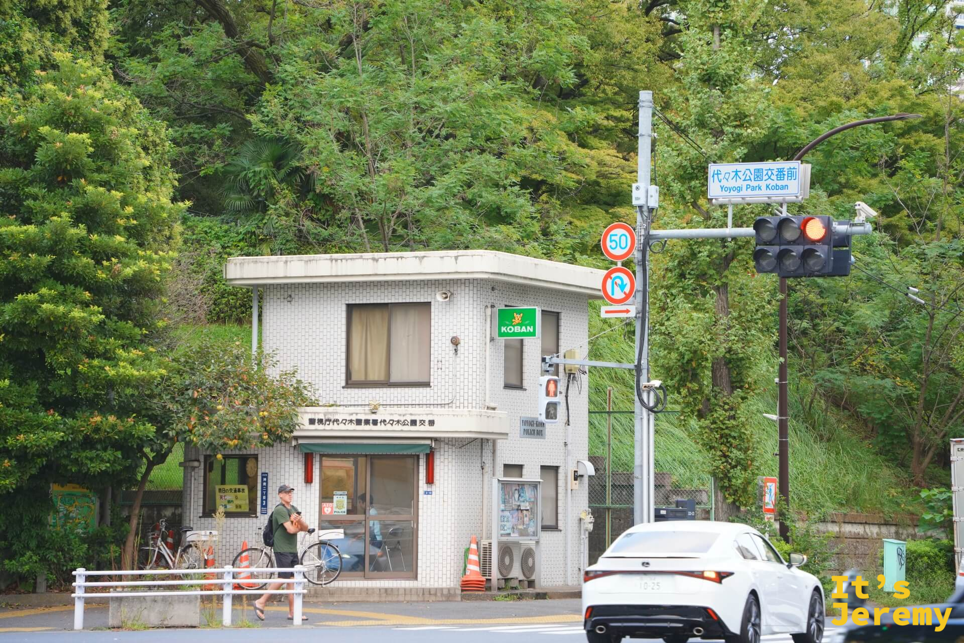 代々木公園交差點,角落是白色交番建築,旁邊路牌寫著「代々木公園交差点」,周圍樹木茂密,白色轎車靜靜停在路邊