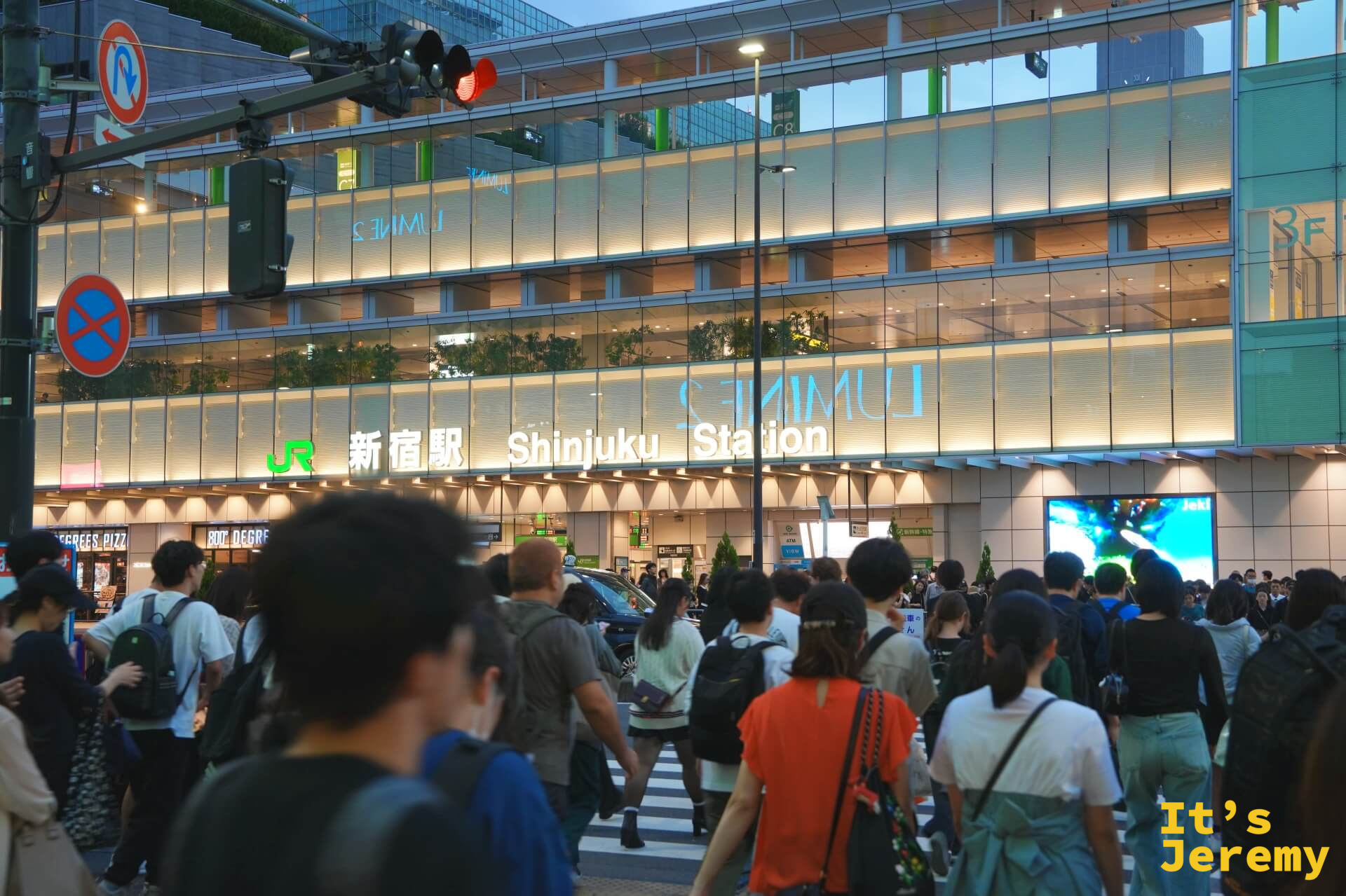 傍晚的 JR 新宿駅南口,LUMINE 玻璃帷幕大樓燈光全亮,「新宿駅 Shinjuku Station」招牌打燈,路口人群正在穿越斑馬線