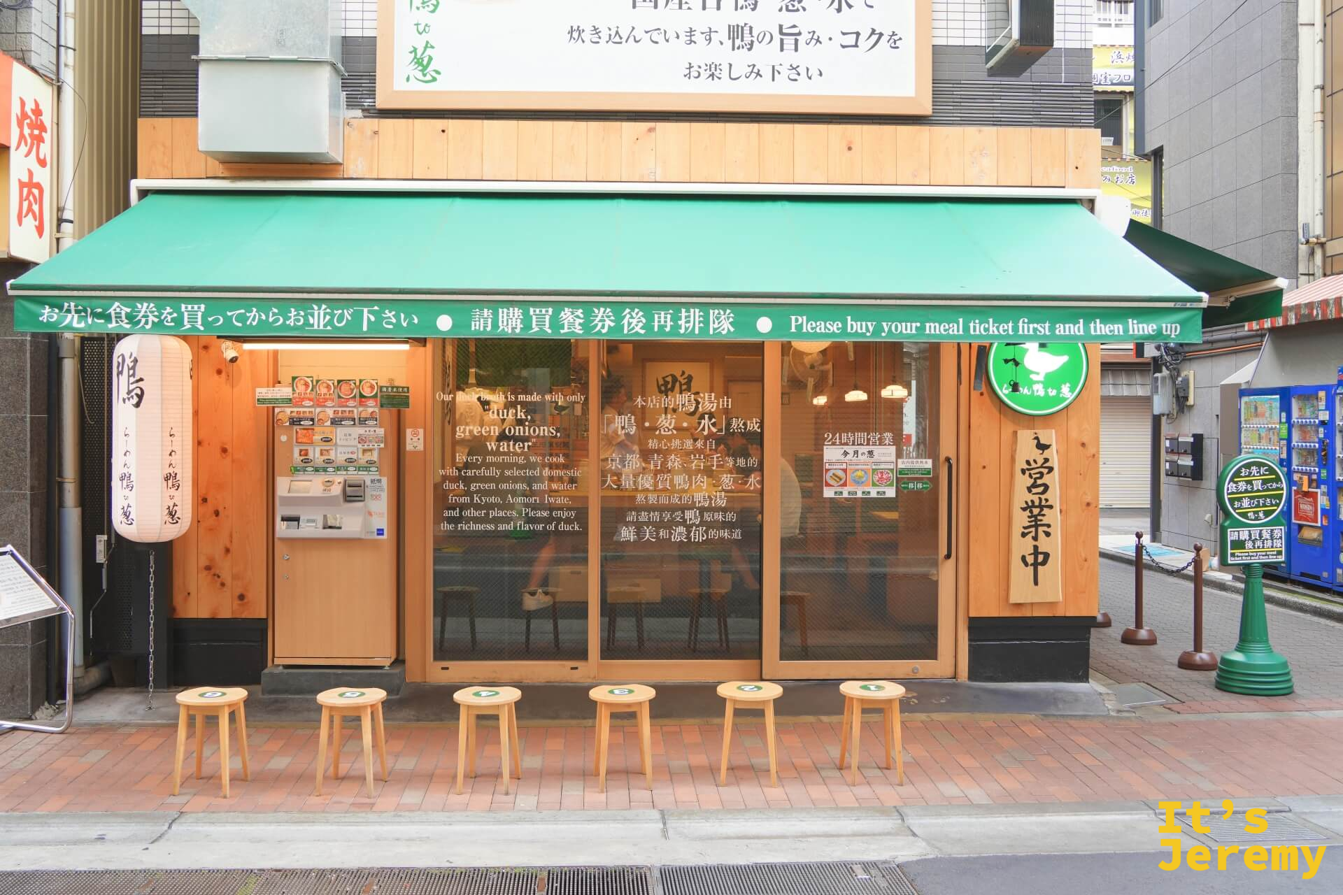 らーめん鴨to葱上野御徒町本店外觀,綠色遮陽棚,門口排隊用的木凳整齊排開,玻璃門上有中日英三語說明