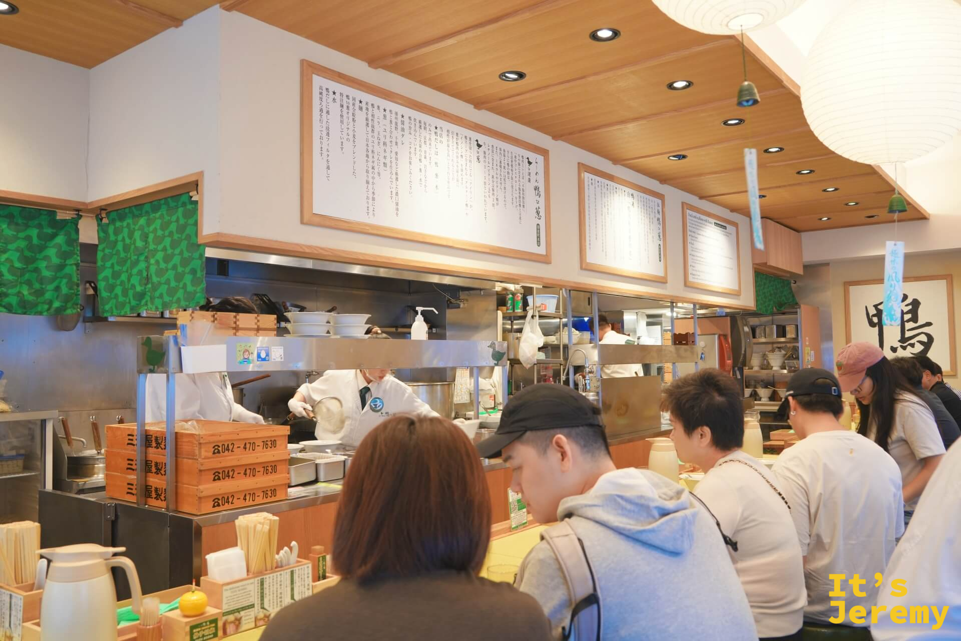 店內吧台座位,多位客人面對廚房坐下,廚師忙碌,牆上說明文字框,右側掛著「鴨」字書法