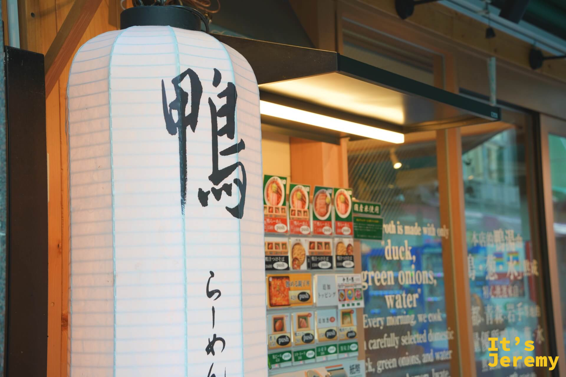 店門口懸掛的白色燈籠,寫著「鴨らーめん」,後方是自動售票機菜單