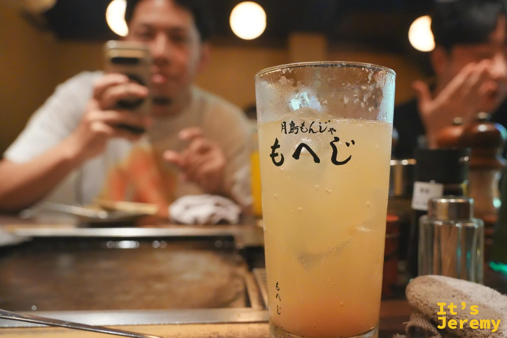 もへじ 店名玻璃杯裝的黃橙色飲品,背景是冒煙的鐵板與食材
