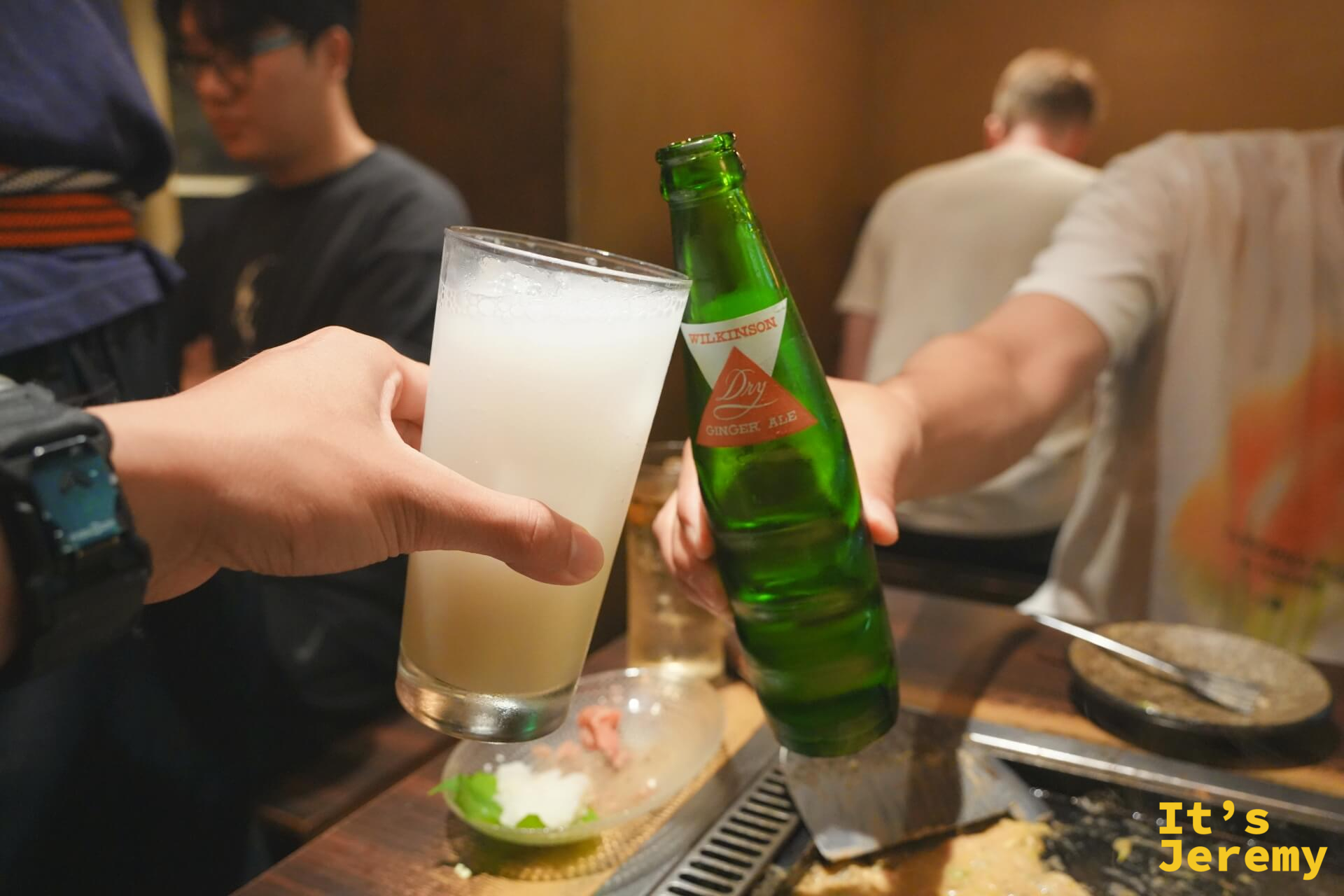 舉起一杯白色混濁飲品,對面是 Wilkinson Dry Ginger Ale 綠色玻璃瓶,周圍是冒著熱氣的鐵板