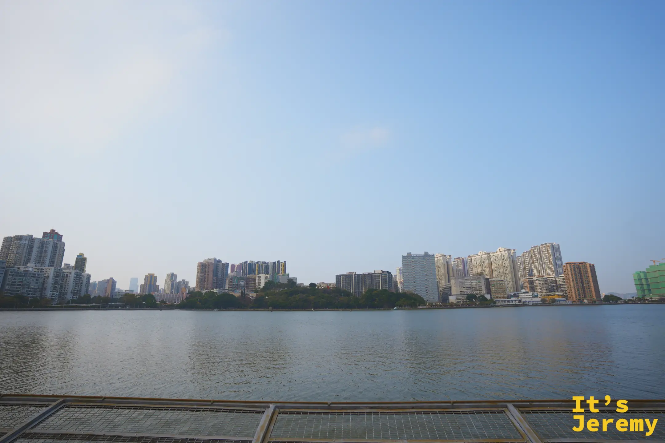 開闊的南灣湖水面，對岸是層疊的住宅高樓與遠山，前方是金屬格柵欄杆