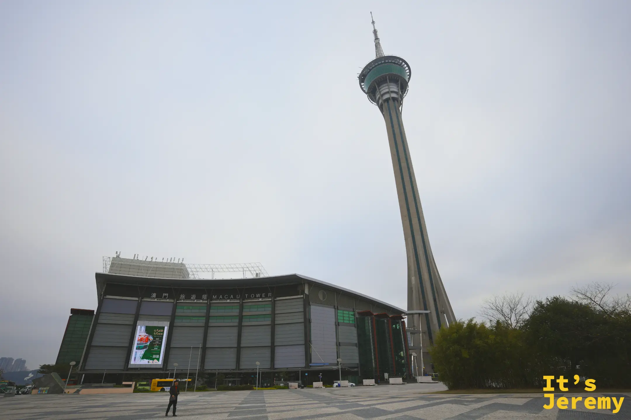 從廣場看澳門塔全景，塔身與基座建築在天空下矗立，前方廣場有行人與黃色巴士