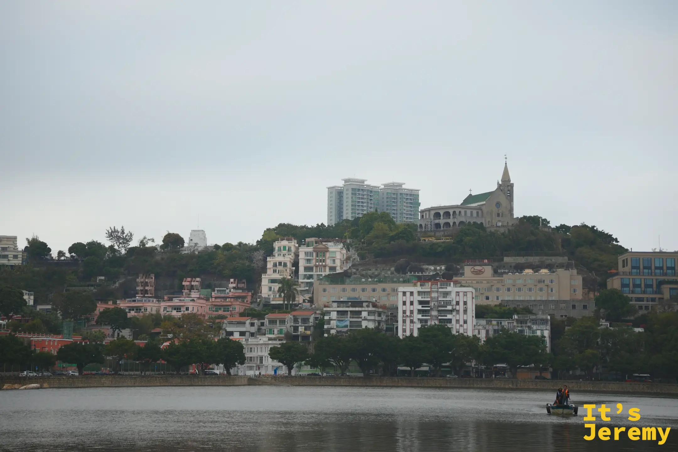 對岸半山上的主教山教堂與尖塔，山坡上是柔和色調的住宅群，水面有一艘小船