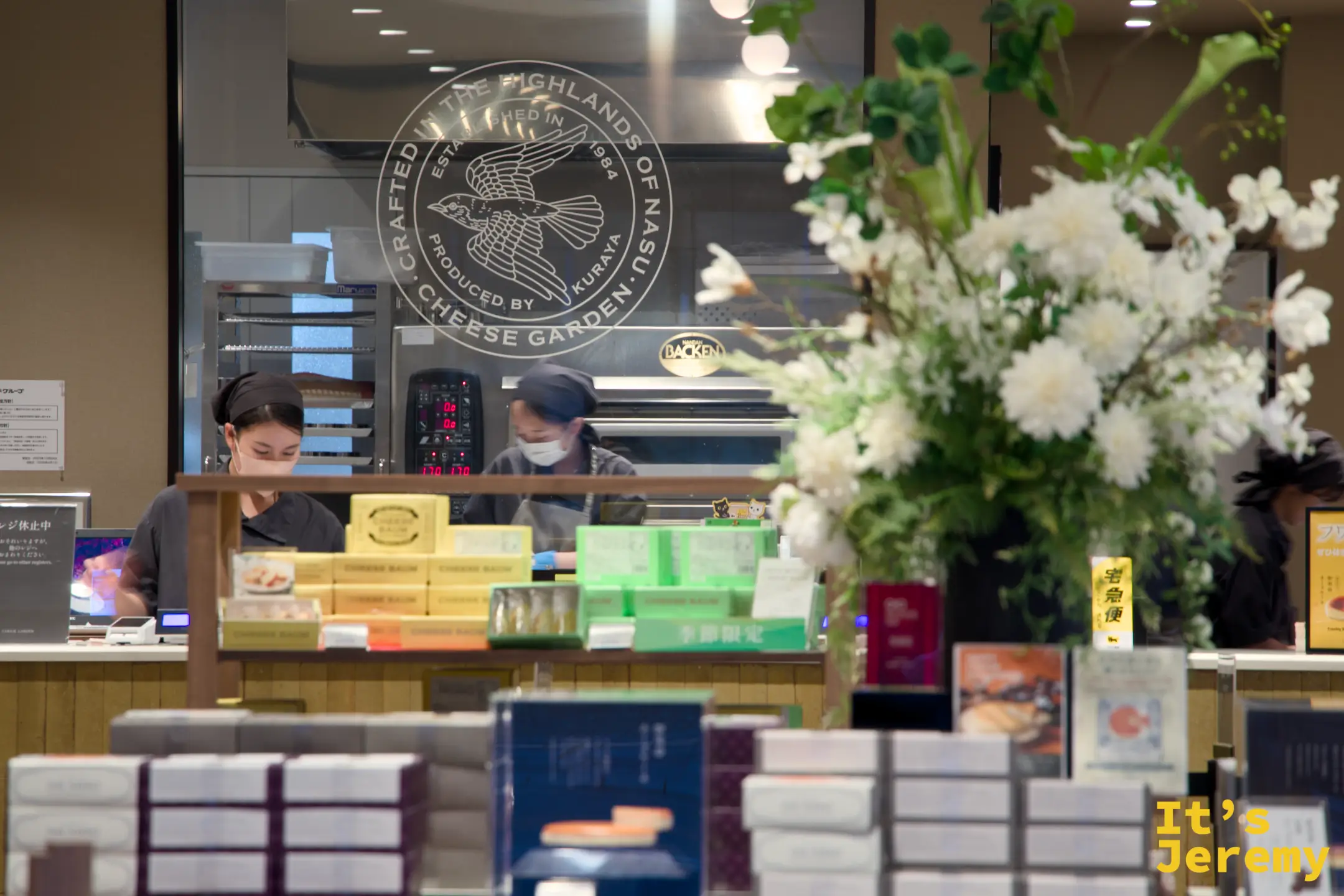 チーズガーデン店内，玻璃透明廚房後工作人員在工作，前方是整齊堆疊的禮盒商品