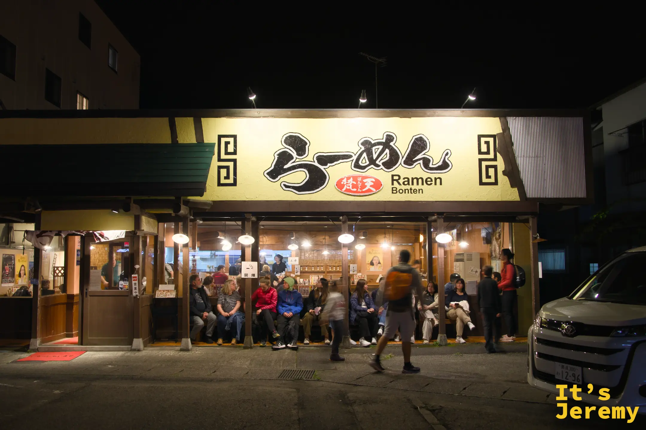 らーめん梵天夜晚外觀，黃色大招牌燈亮，店外長椅坐滿等位的旅人