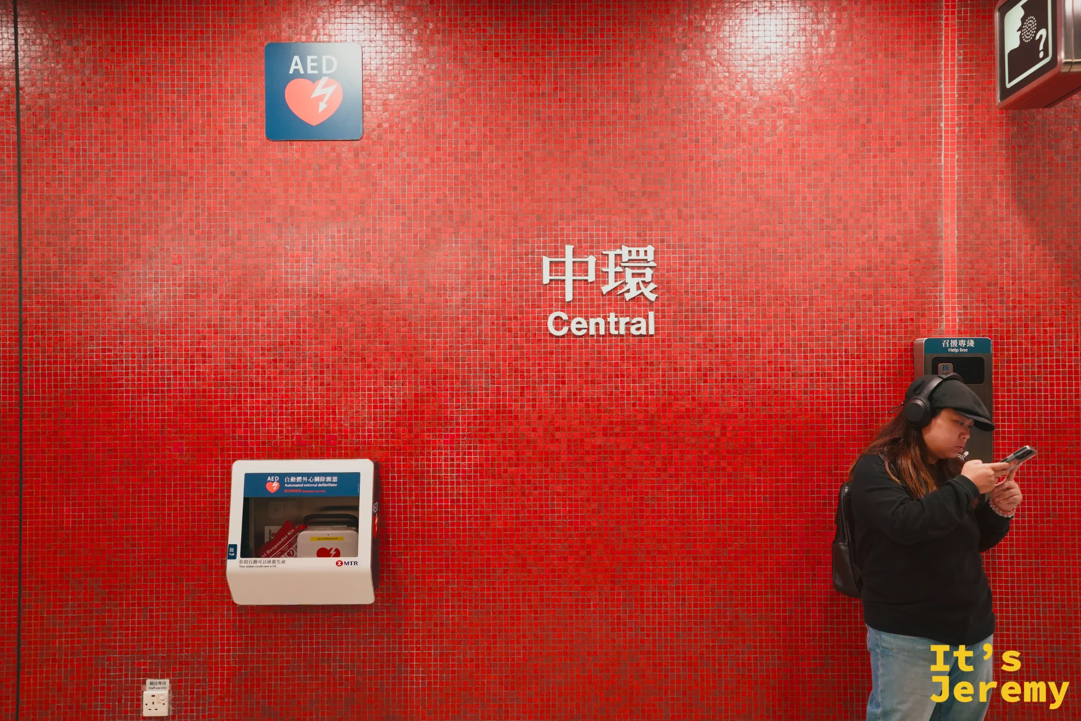 港鐵中環站的紅磚色馬賽克牆面，白字寫著「中環 Central」，一旁有 AED 與乘客
