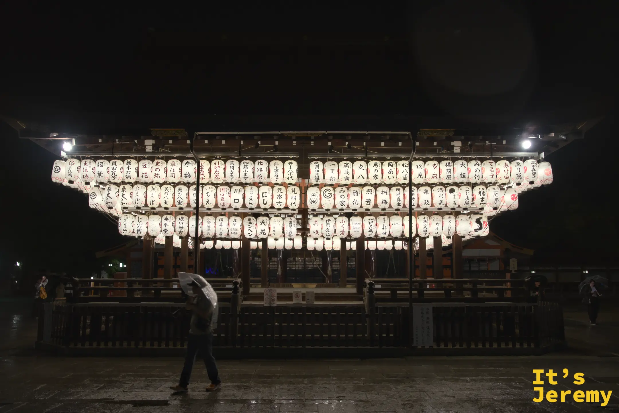 舞殿提燈全景，有撐傘遊客路過，雨夜氛圍最濃