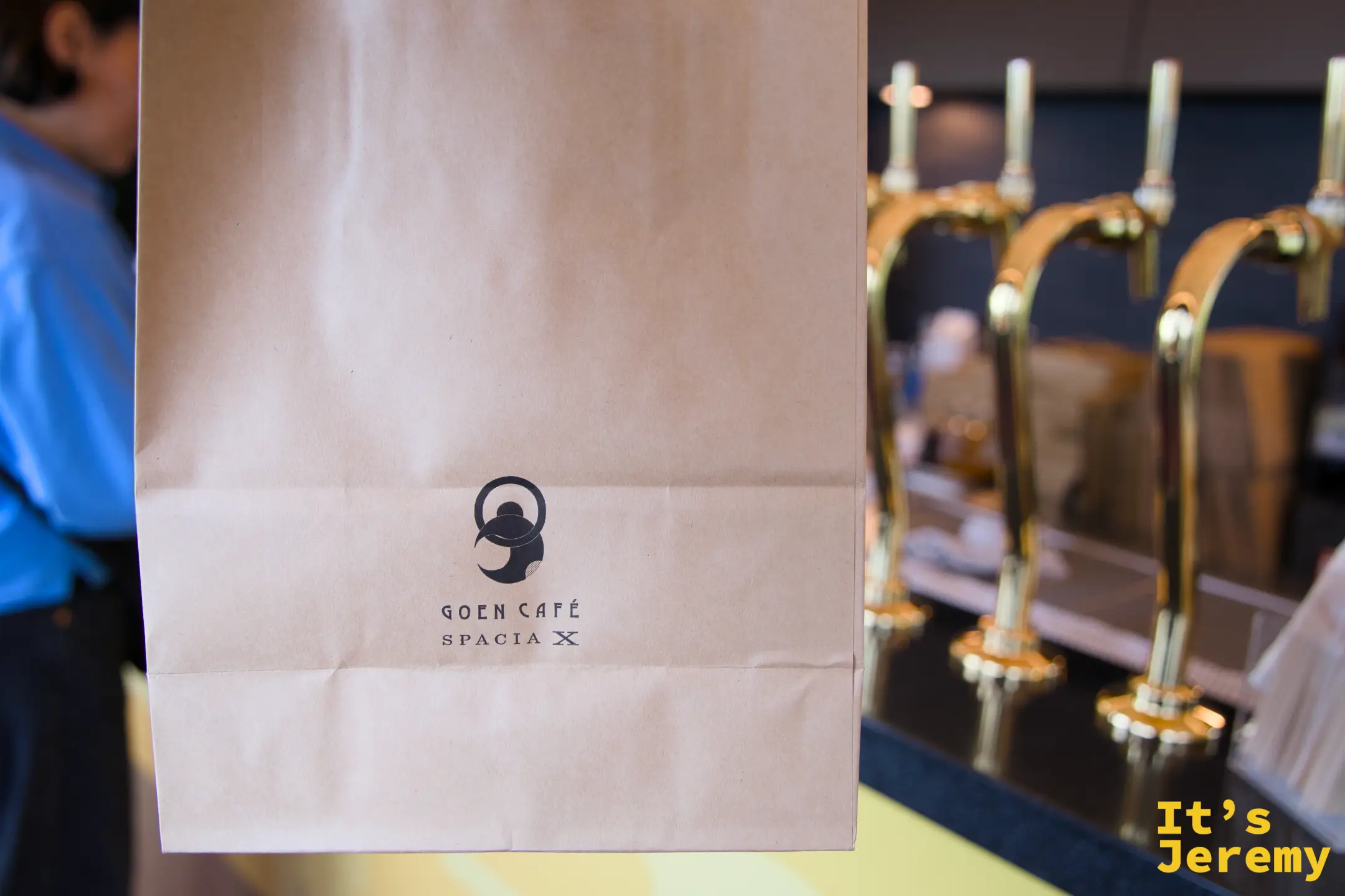 GOEN CAFÉ SPACIA X 牛皮紙袋正面，印有 GOEN CAFÉ 壺形 logo 和 SPACIA X 字樣，後方是金色生啤酒龍頭