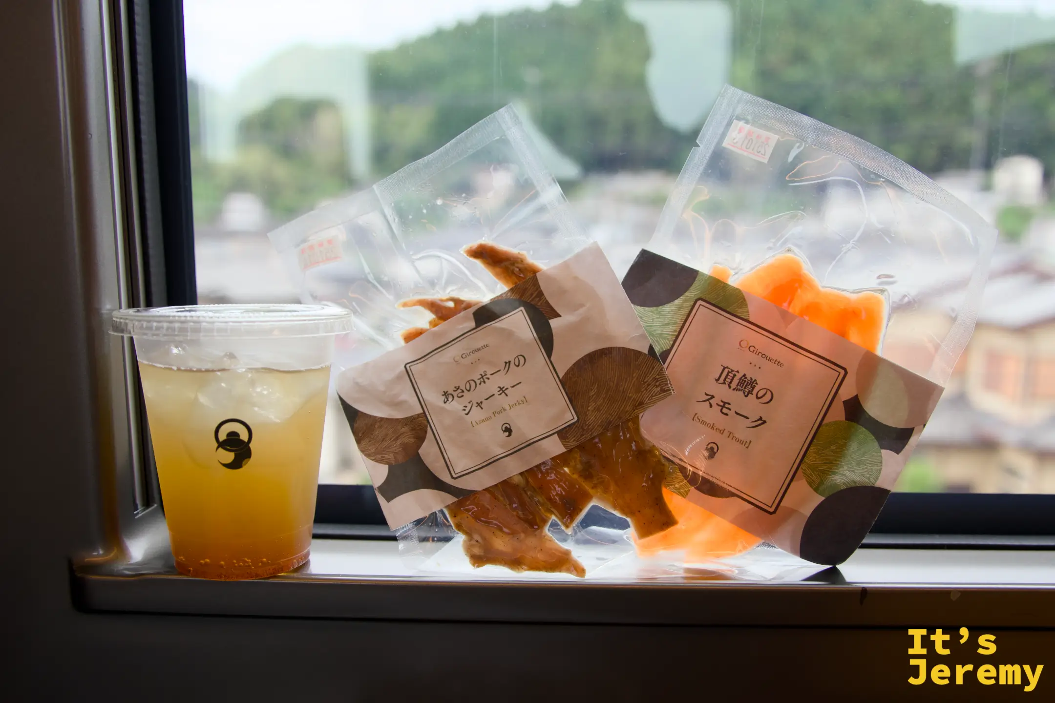 SPACIA X 車窗旁，ニッコーラ飲料杯、あさのポークのジャーキー、頂鱒のスモーク三件並排，窗外是山區景色