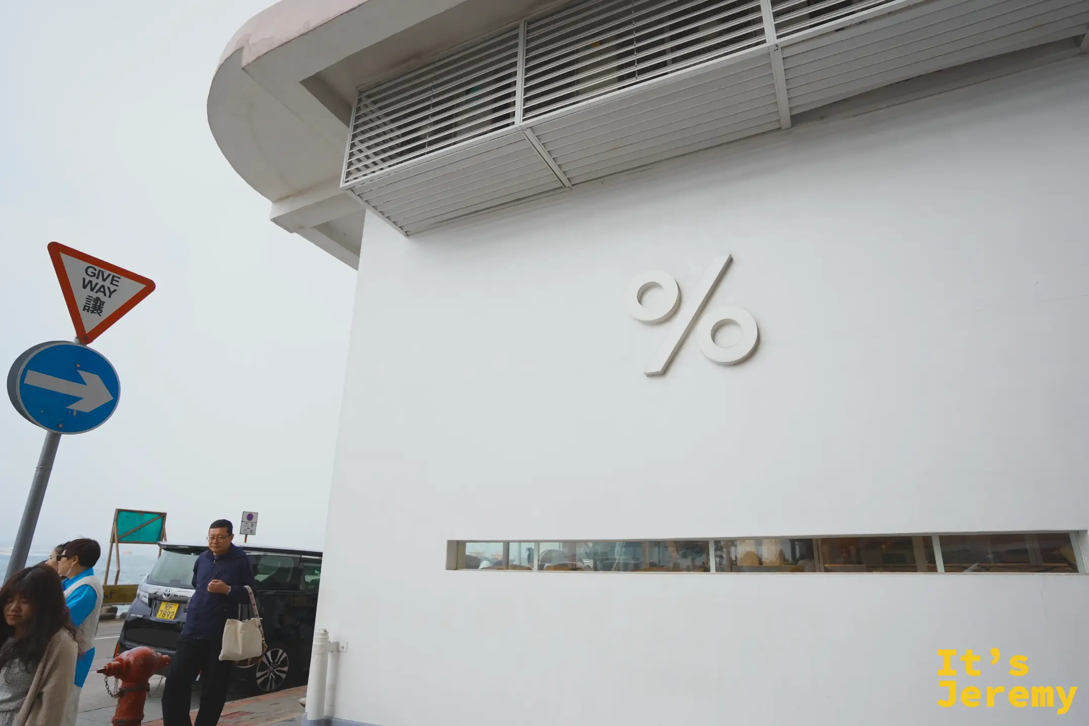 %Arabica 白色建築牆面上的立體「%」logo 特寫，上方是白色通風百葉，下方橫向落地窗，一名男子從樓下走過