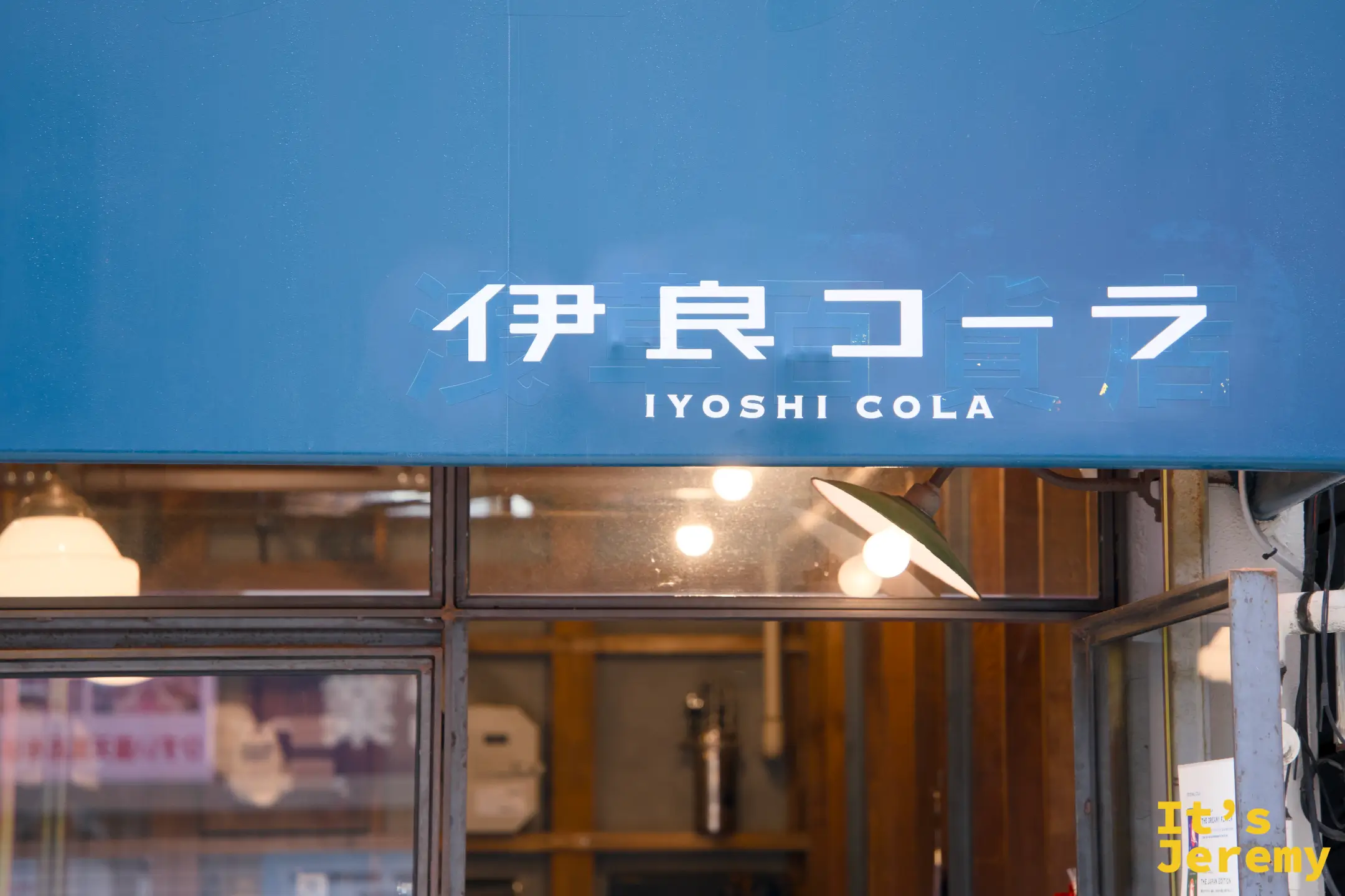 青藍色遮陽棚近景，「伊良コーラ IYOSHI COLA」白字清楚