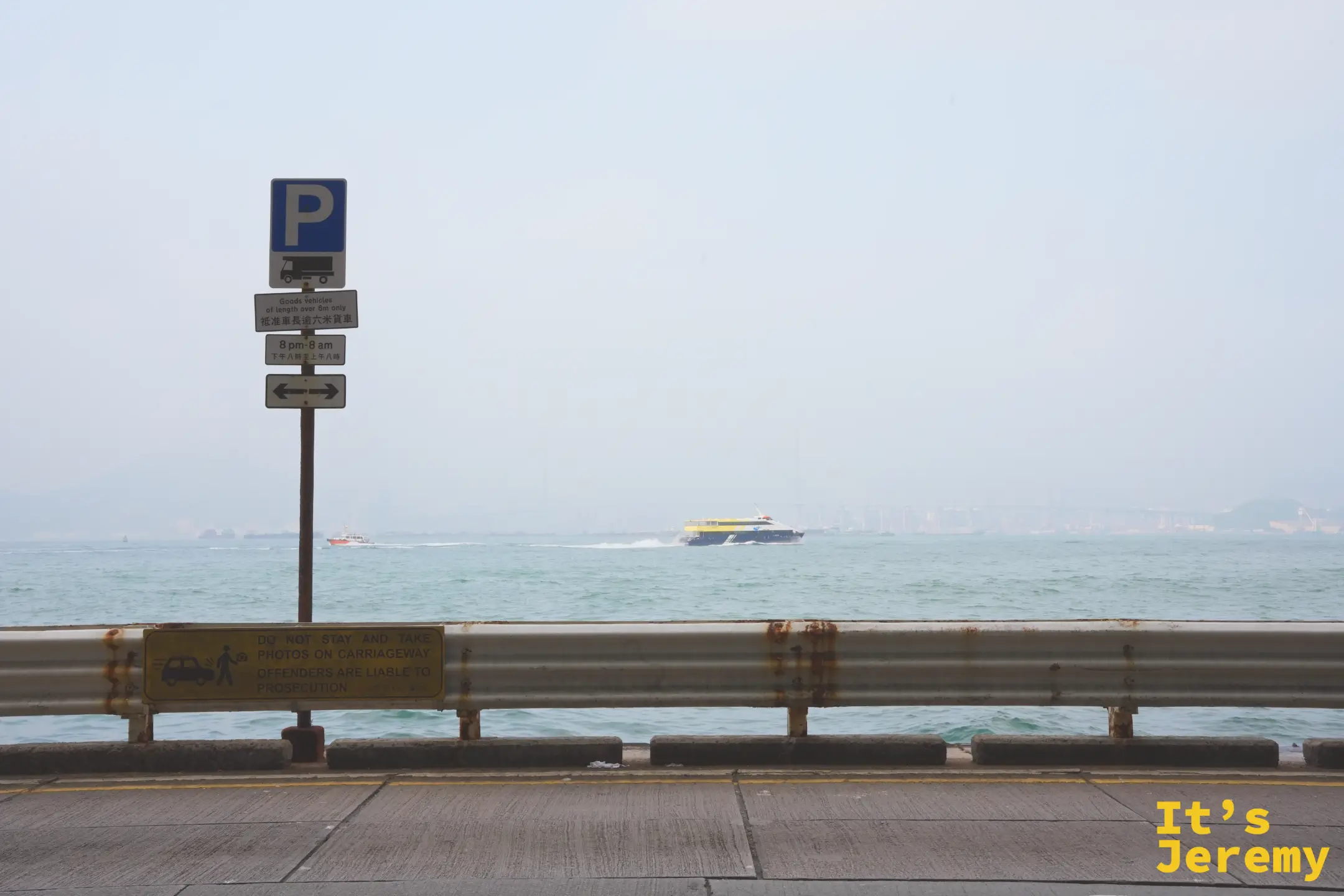 堅尼地城海旁的護欄與交通告示牌，寫著「DO NOT STAY AND TAKE PHOTOS ON CARRIAGEWAY」，海面上有渡輪與橋樑剪影