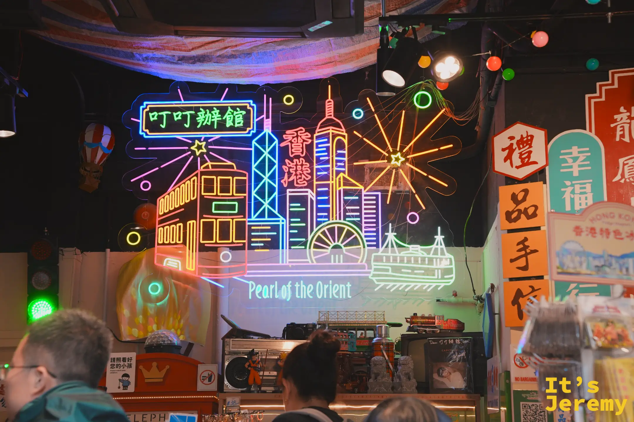 叮叮辦館手信店內，牆上霓虹燈勾勒出香港天際線、叮叮電車與 Pearl of the Orient 字樣，架上有懷舊小物與禮品招牌