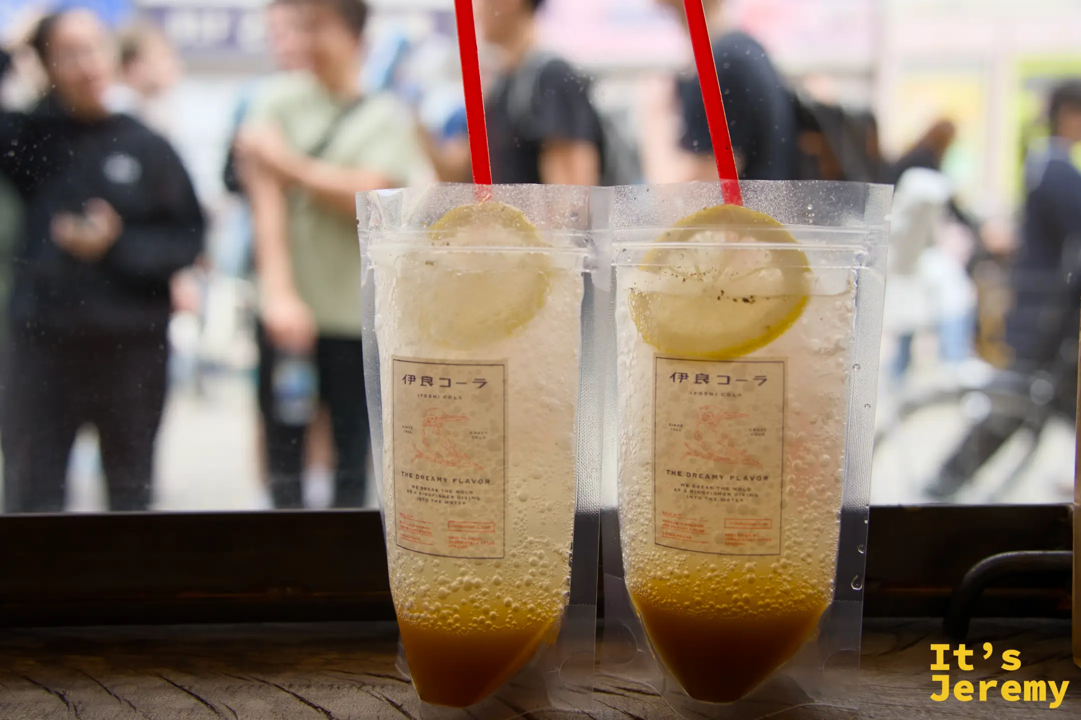 兩杯伊良コーラ 裝在透明杯裡，插著紅色吸管、放上檸檬片，杯身印有品牌字樣，背景是浅草街頭人群
