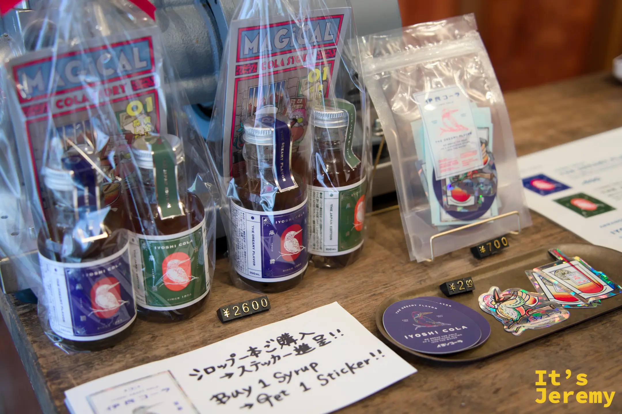 收銀台旁的糖漿套組、貼紙與周邊商品，手寫牌寫著「Buy 1 Syrup → Get 1 Sticker!!」