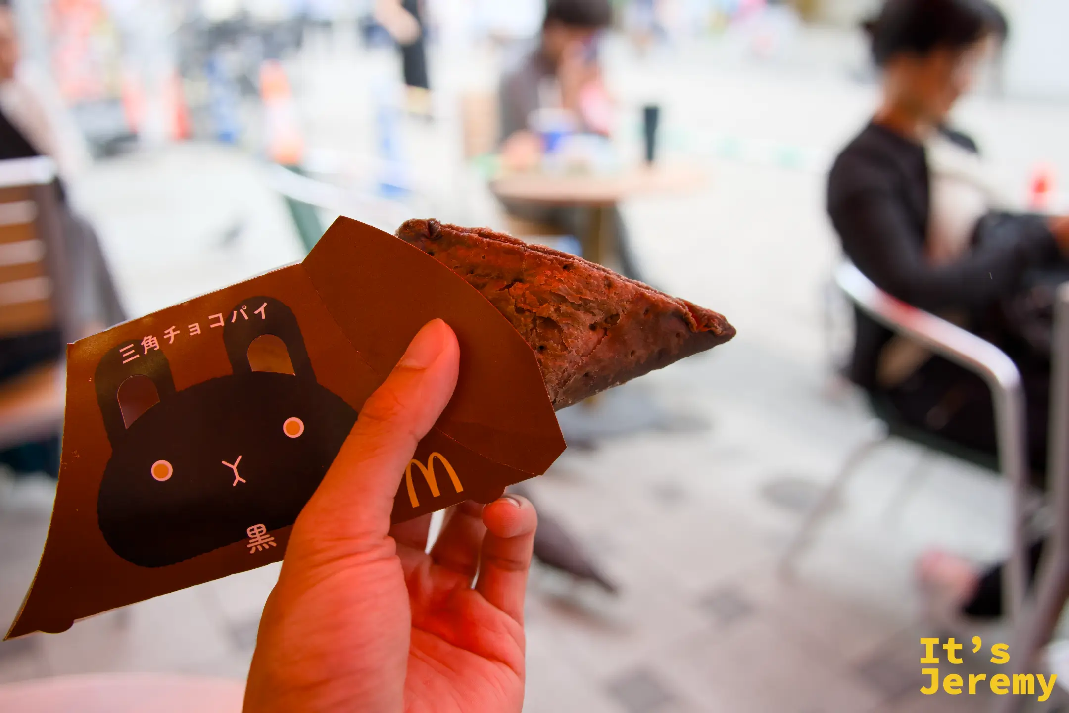 手持三角チョコパイ(黒),麥當勞紙盒上印著兔子圖案與「黒」字,背景是麥當勞室內座位區