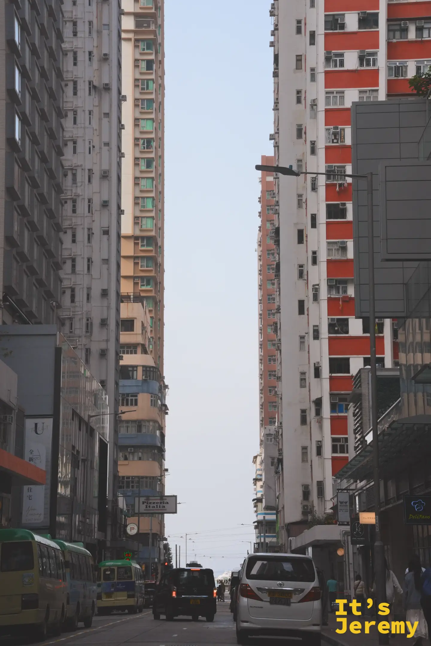 堅尼地城街道景，灰濛濛的天色下小巴、計程車與行人並行，遠方掛著 Pizzeria Italia 招牌