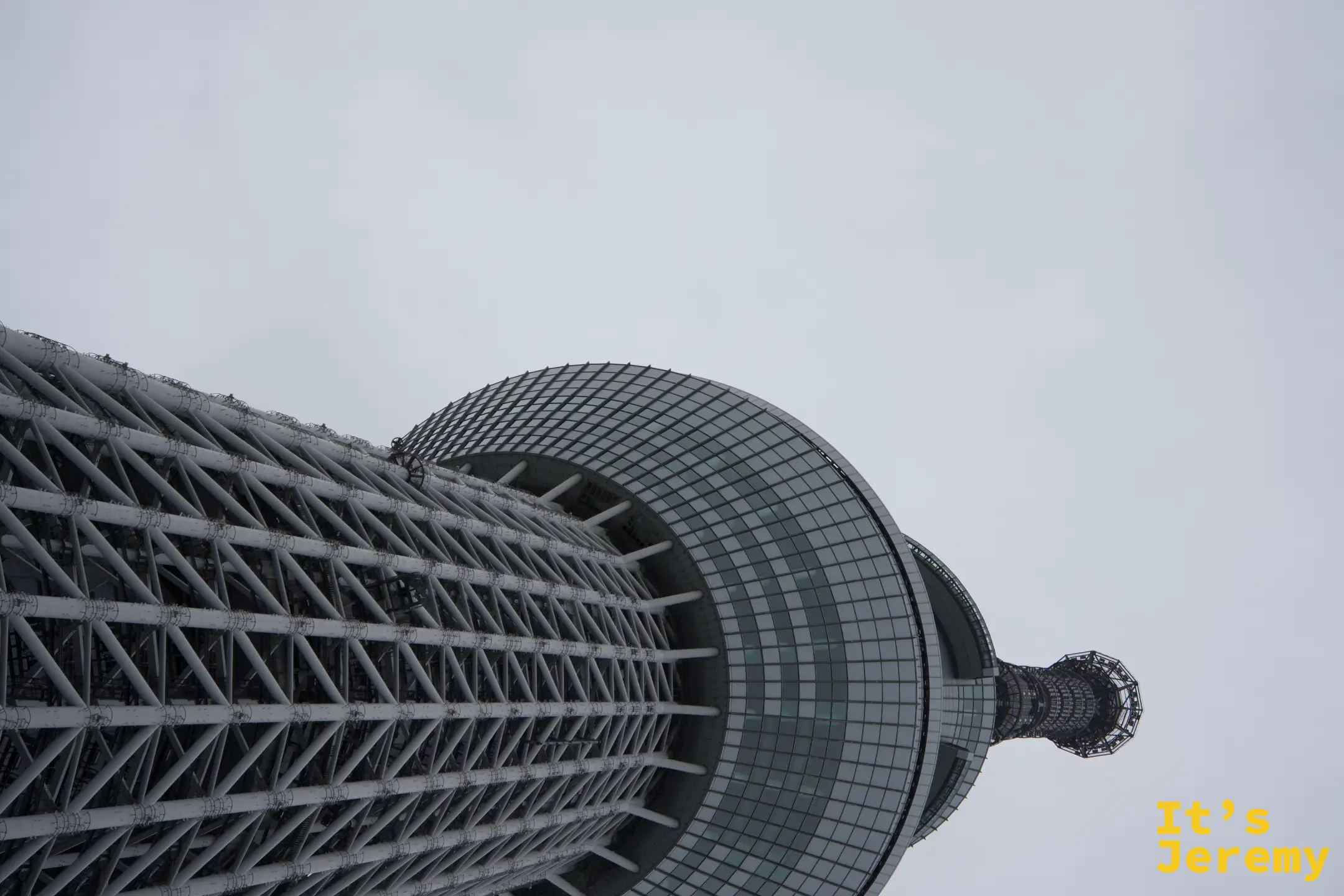 仰望晴空塔塔身,鋼骨菱格結構向上延伸,天望台圓球清晰可見,灰色天空為背景