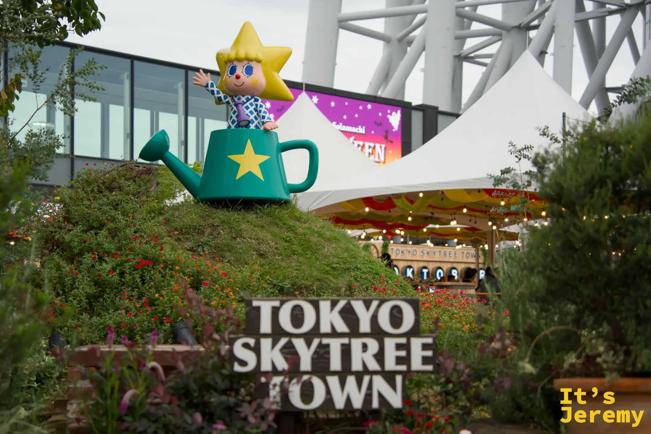 Tokyo Skytree Town 廣場入口,吉祥物坐在澆花壺造型座上,周圍花圃盛開,招牌清晰可見