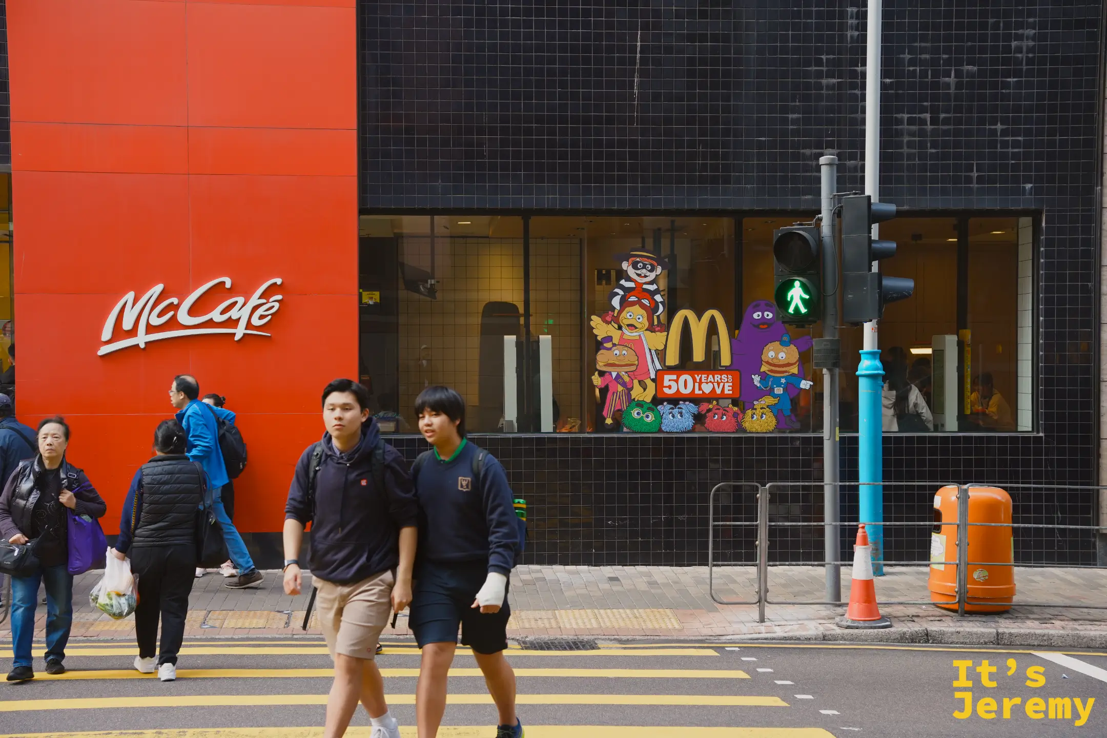 堅尼地城街角的 McCafé，黑磚外牆與紅色招牌，落地窗貼著麥當勞 50 週年的角色圖案，斑馬線上有兩名學生走過