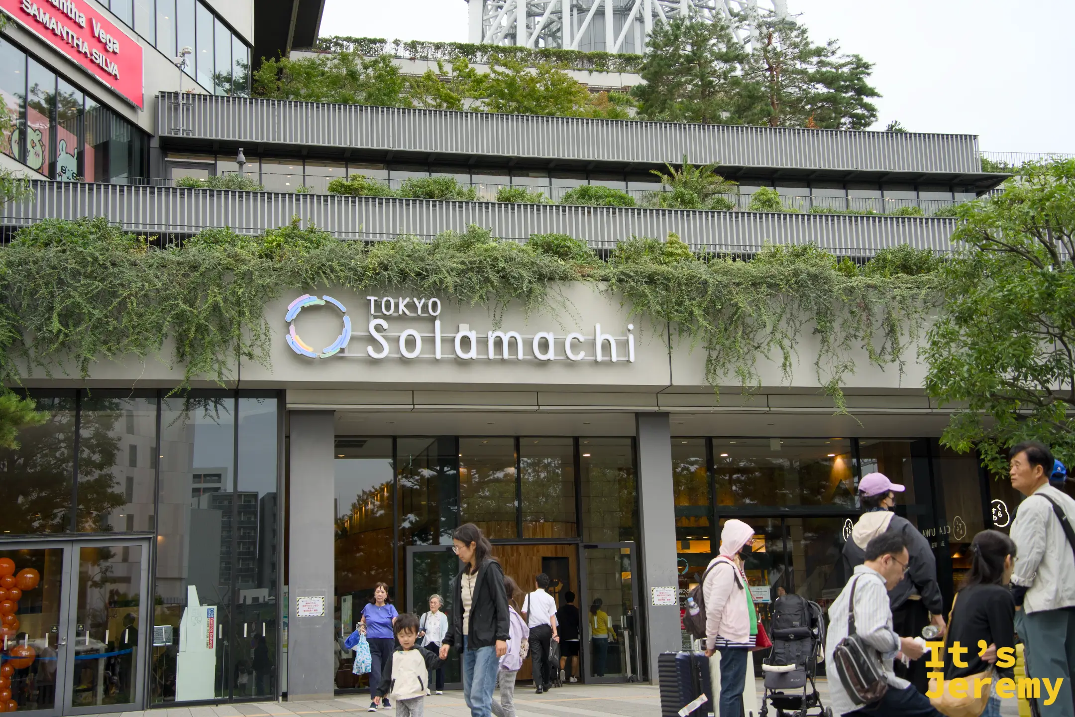 Tokyo Solamachi 商場正面入口,藍色細字 logo 清晰,玻璃帷幕建築頂部種有植栽,行人進出穿梭