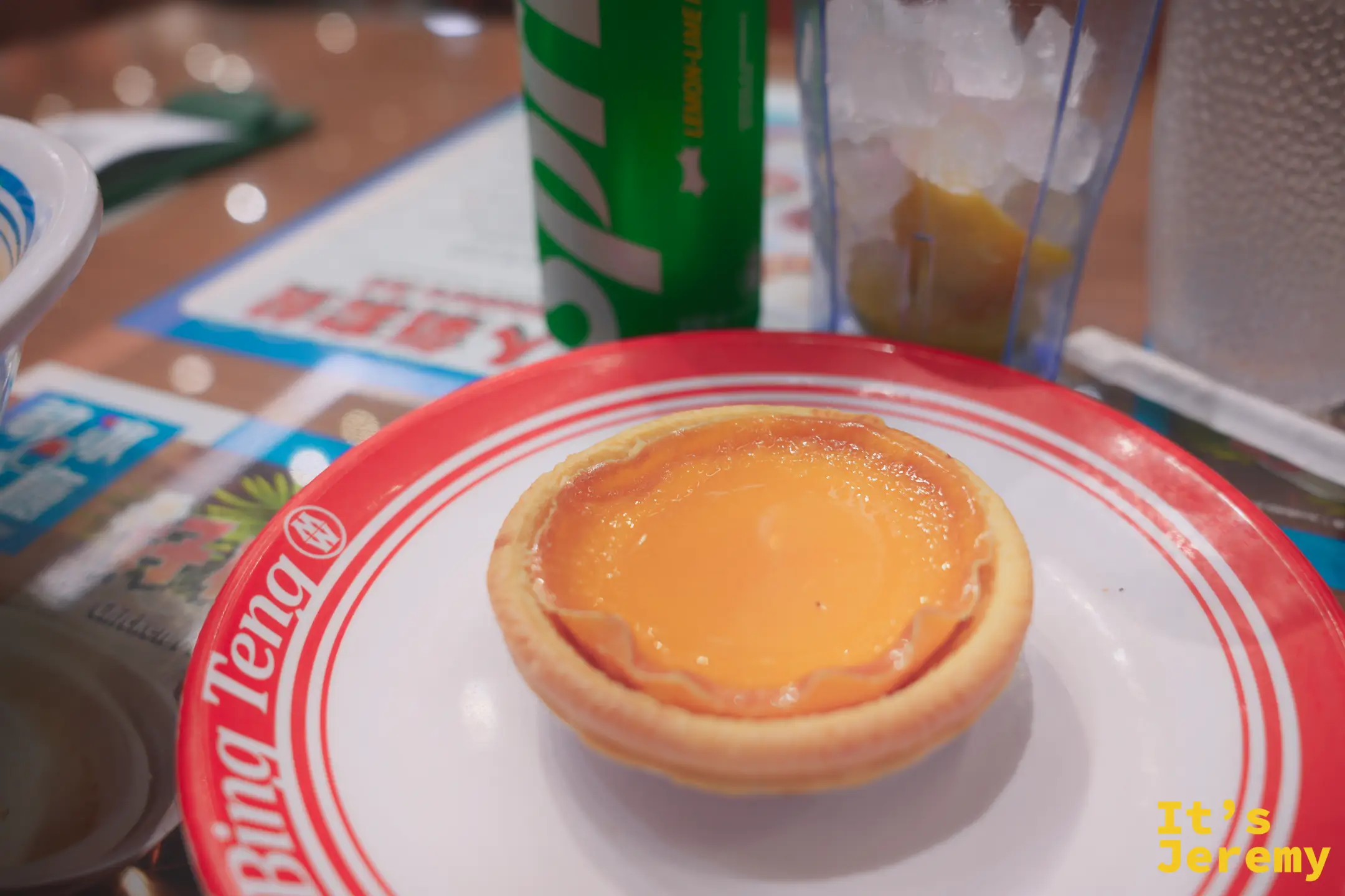 橘色條紋「Bing Teng」瓷盤上放著一顆金黃酥皮蛋撻，中央蛋漿飽滿滑嫩，後方是綠色雪碧罐與一杯加冰的鹹檸檬飲料