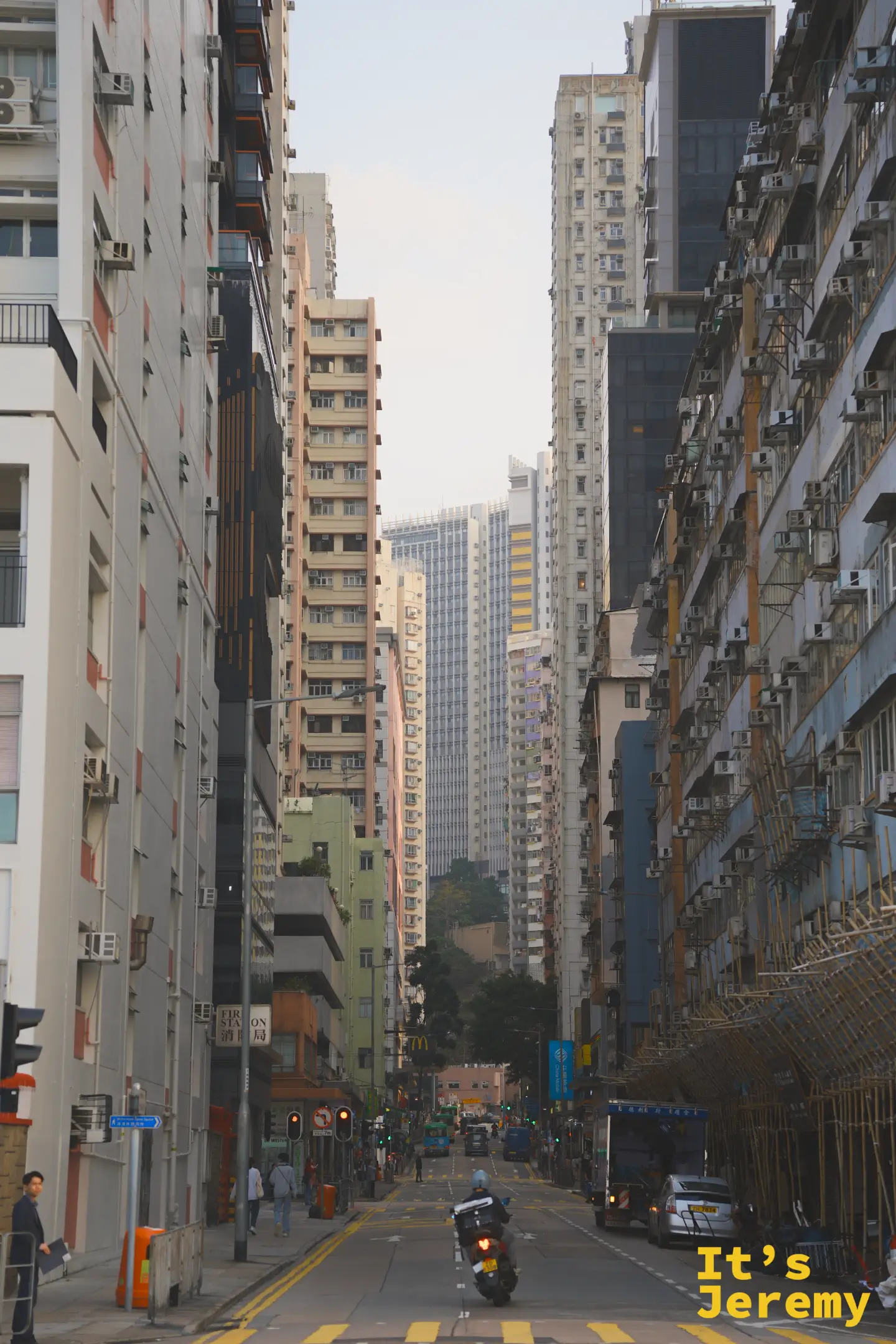 從街道望向遠方的長坡，兩旁是層層疊高的住宅大樓，路上有機車、貨車與棚架，遠處立著消防局招牌