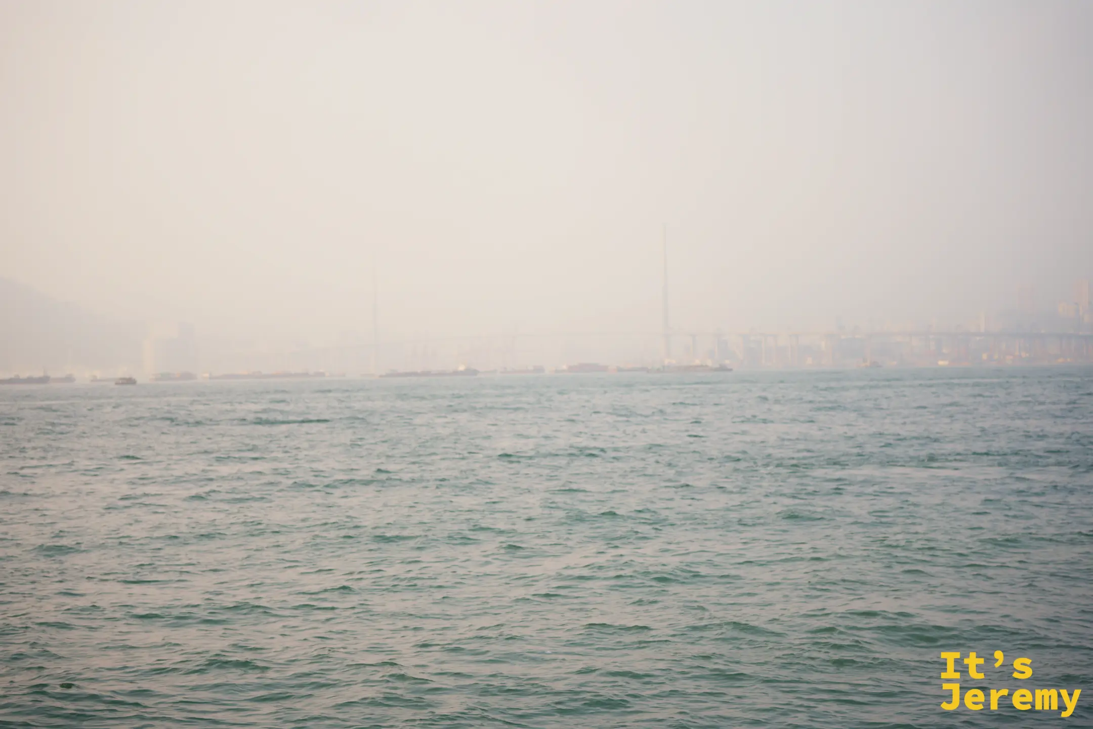 霧濛濛的維多利亞港海面，遠方的橋樑與高樓化成淡淡剪影，海水呈帶灰的青綠色