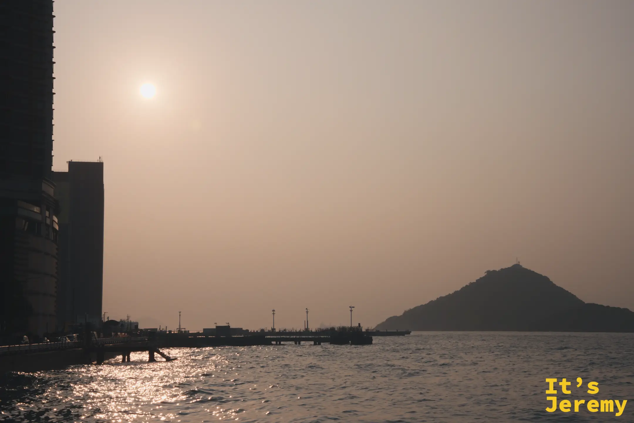 堅尼地城海旁的黃昏，夕陽掛在左側高樓邊，把海面鋪成金黃色，棧橋與遠方小山化成深色剪影