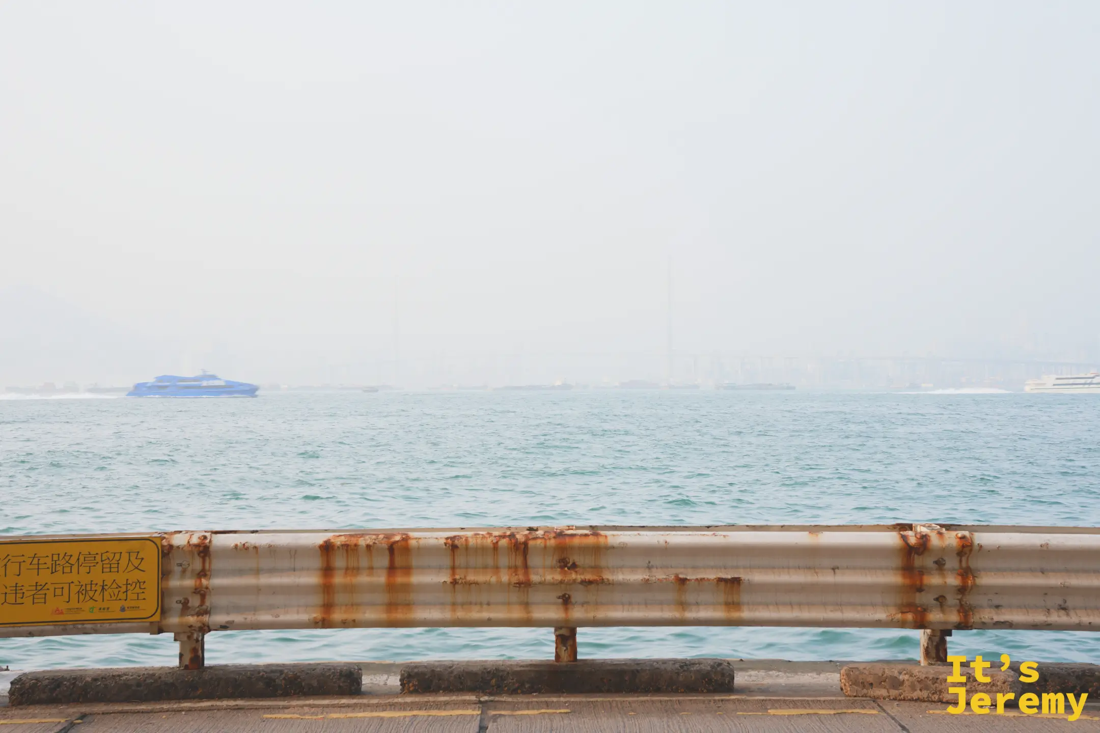 堅尼地城海旁，藍色高速渡輪駛過灰藍海面，遠方是霧中的橋樑與貨櫃碼頭，前景是生鏽的護欄
