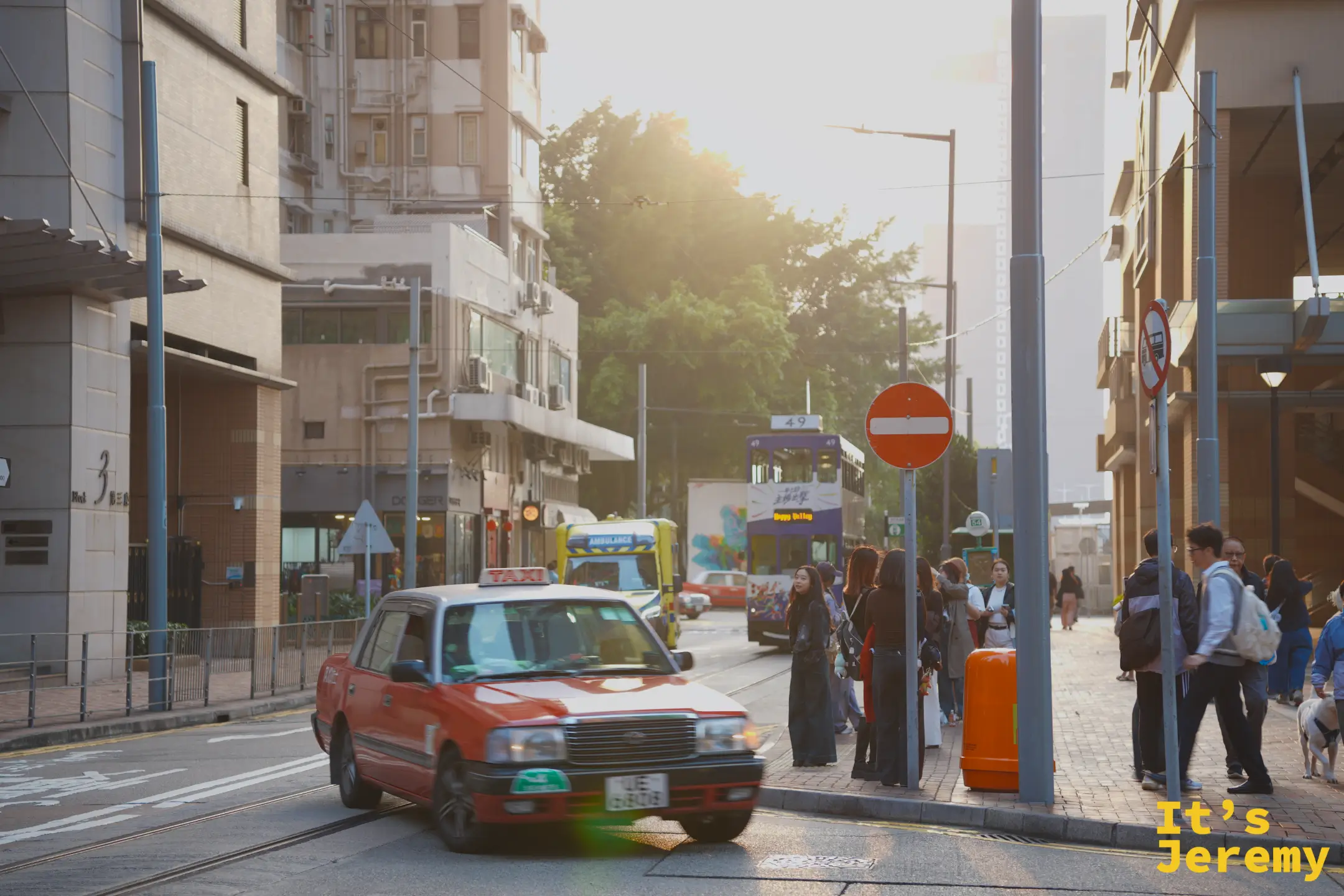 堅尼地城街角，紅色計程車停在路邊，背後陽光從街尾灑下形成光暈，人們在電車站等車，還有一隻狗