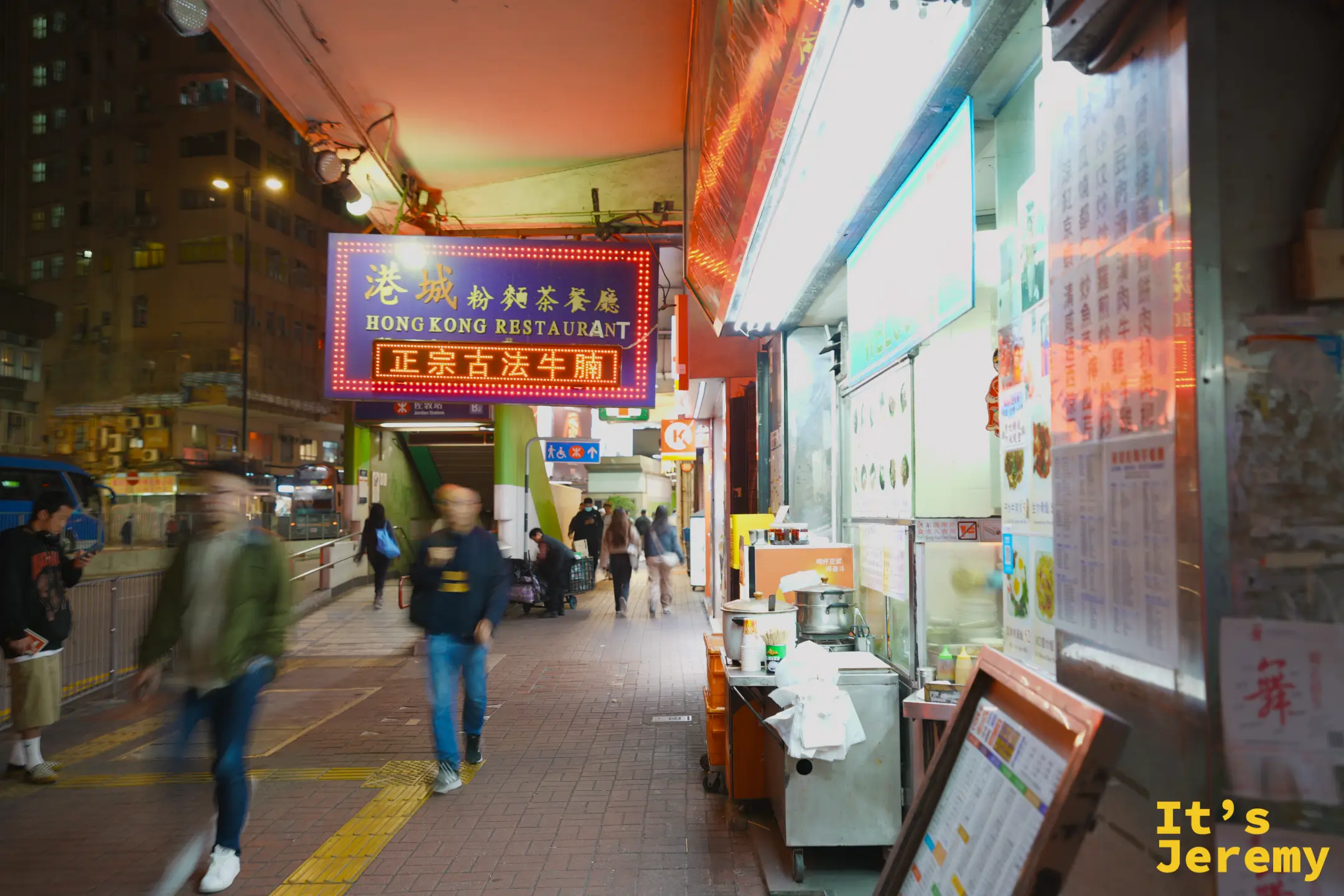 佐敦港鐵站旁的騎樓通道，港城粉麵茶餐廳的霓虹招牌與「正宗古法牛腩」燈牌亮著，人們往來穿梭，一旁是貼滿菜單的檔口