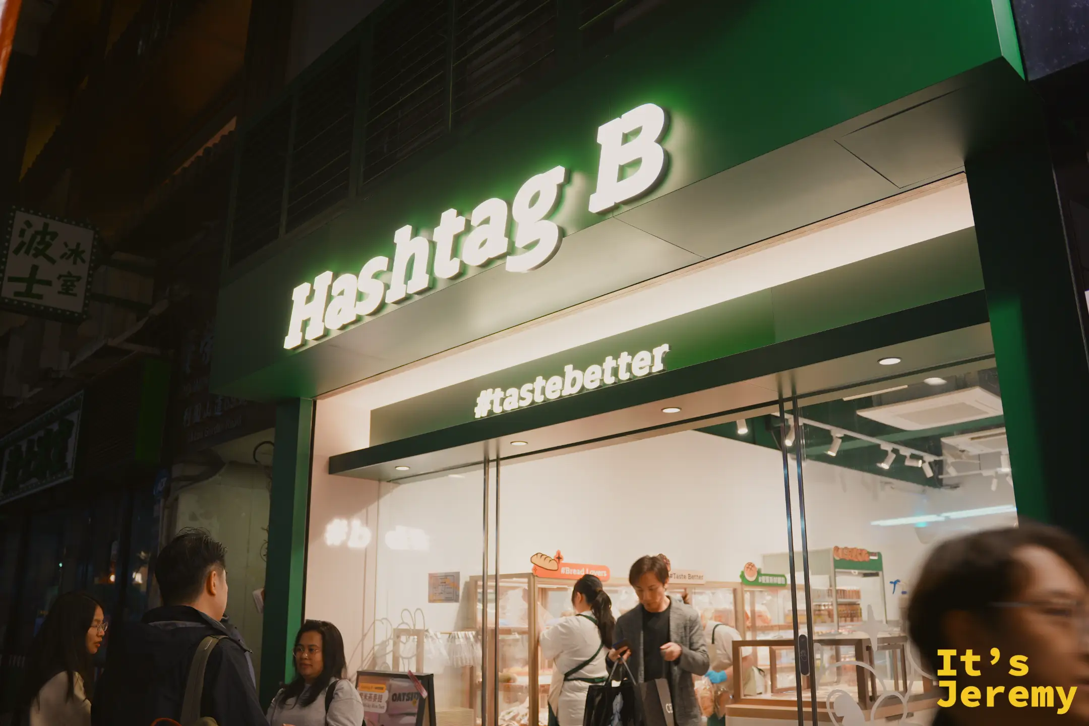 夜裡的 Hashtag B 銅鑼灣店門面，墨綠色招牌上白色燈字「Hashtag B」與「#tastebetter」，玻璃櫥窗內是明亮的烘焙空間