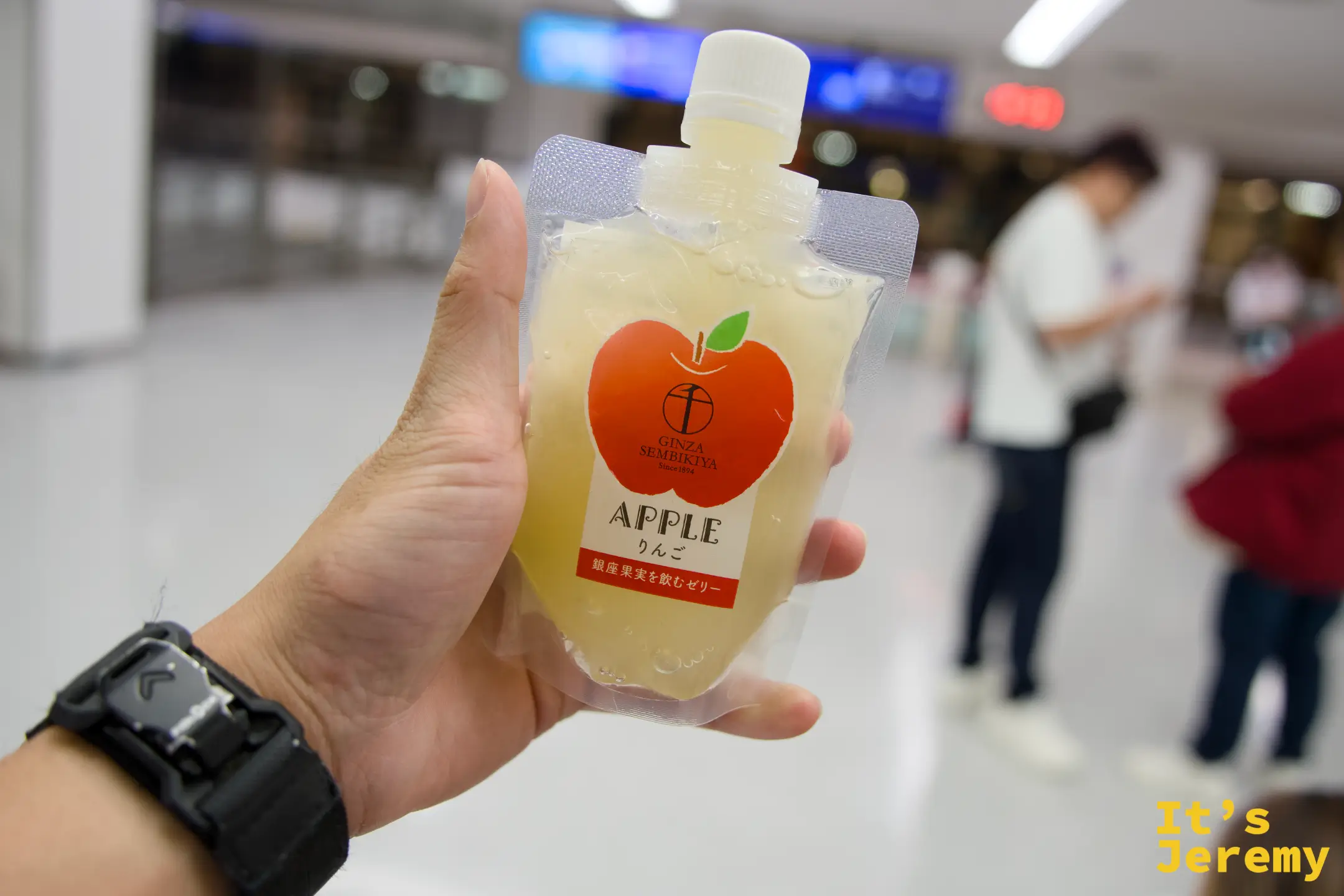 機場大廳,手持 Ginza Sembikiya 蘋果口味袋裝果凍,包裝印有蘋果圖案與品牌 logo,旅客往來背景