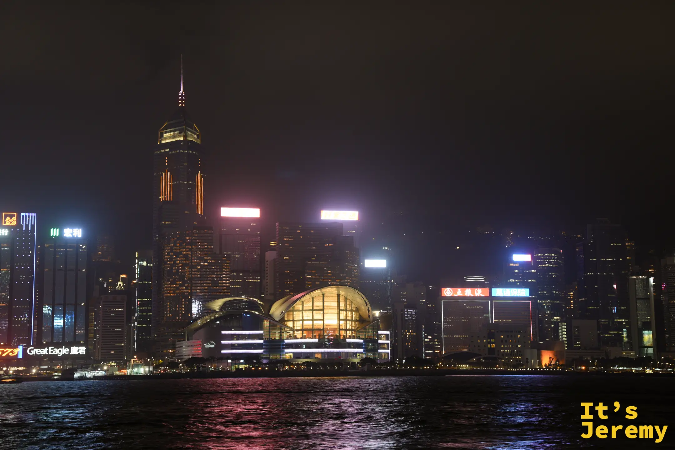 維多利亞港對岸的香港會議展覽中心夜景，波浪般起伏的金屬屋頂被暖黃燈光打亮，倒映在深色海面上，背後是港島高樓